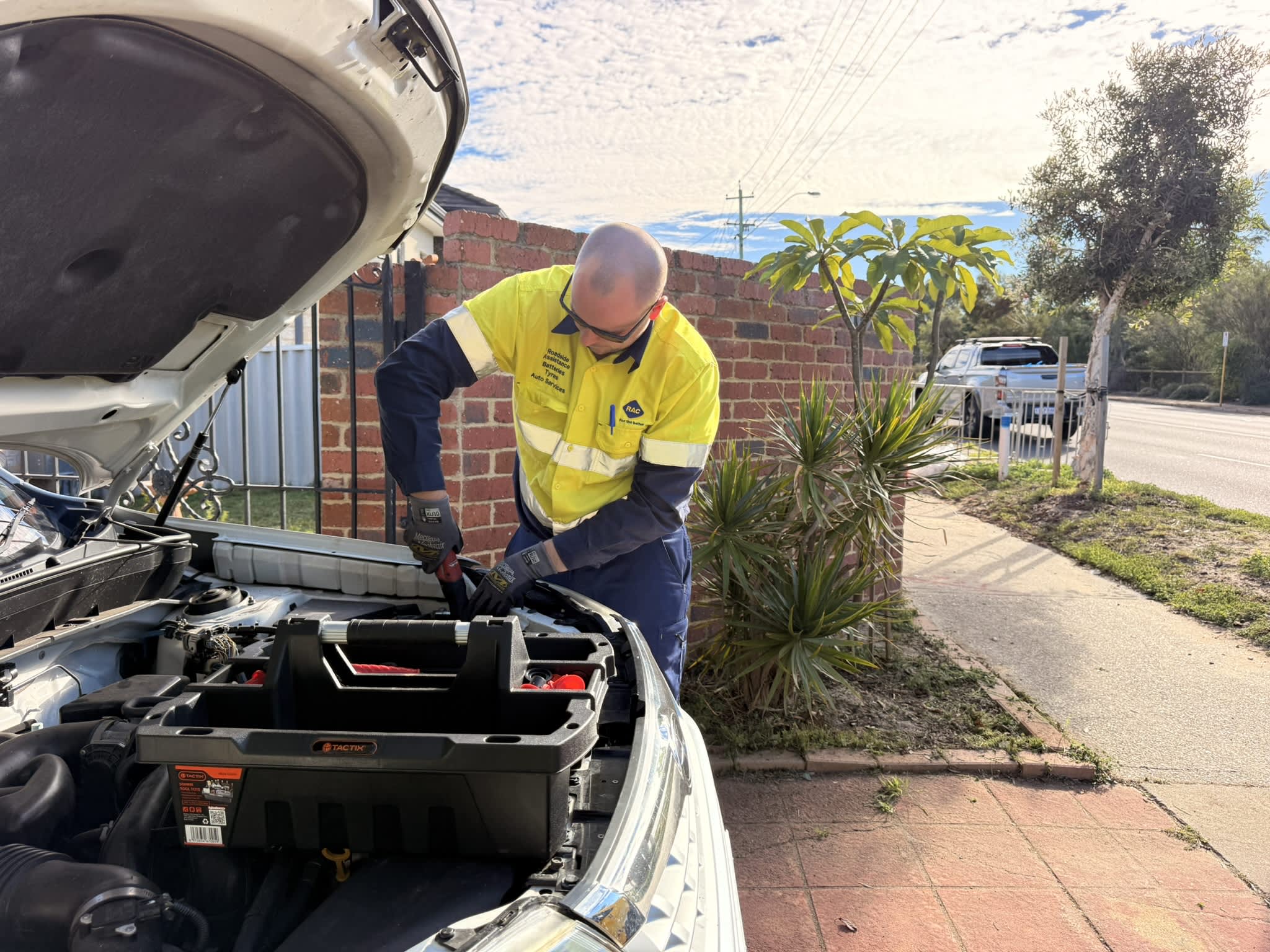 An RAC roadside assistance technician in a high-visibility yellow and navy uniform works under the open hood of a white car parked on a suburban driveway. He wears protective glasses and gloves while using a tool near the car battery. A black plastic tool caddy sits balanced on the engine bay in the foreground. In the background, a brick wall, small trees, and a silver pickup truck parked on the street are visible under a blue sky with scattered clouds.