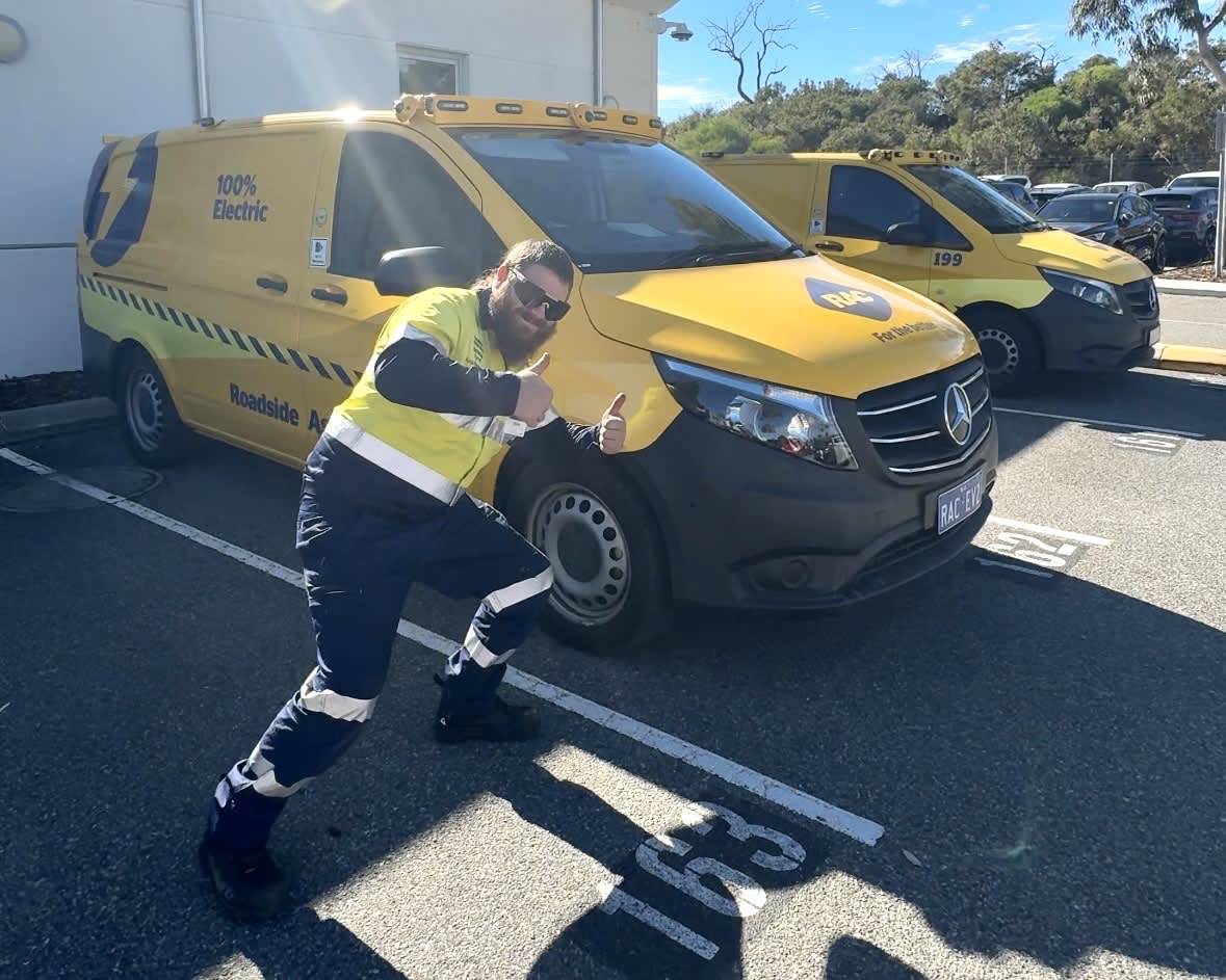 An RAC roadside assistance technician with a beard and sunglasses poses dynamically with double thumbs-up in front of a yellow Mercedes-Benz electric patrol van. He is wearing a high-visibility yellow vest over a navy uniform with reflective strips. The van features a large blue lightning bolt logo on the back side and text reading "100% Electric" and "Roadside Assistance". It is parked in a numbered parking space on an asphalt lot next to another yellow service van under a clear, sunny sky.