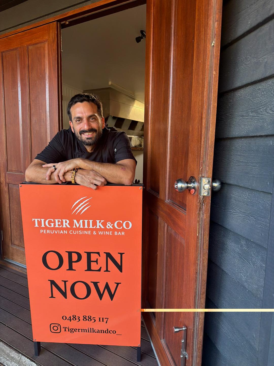 Chef and owner standing out front of restaurant in black clothing with Open Now sign in front of him