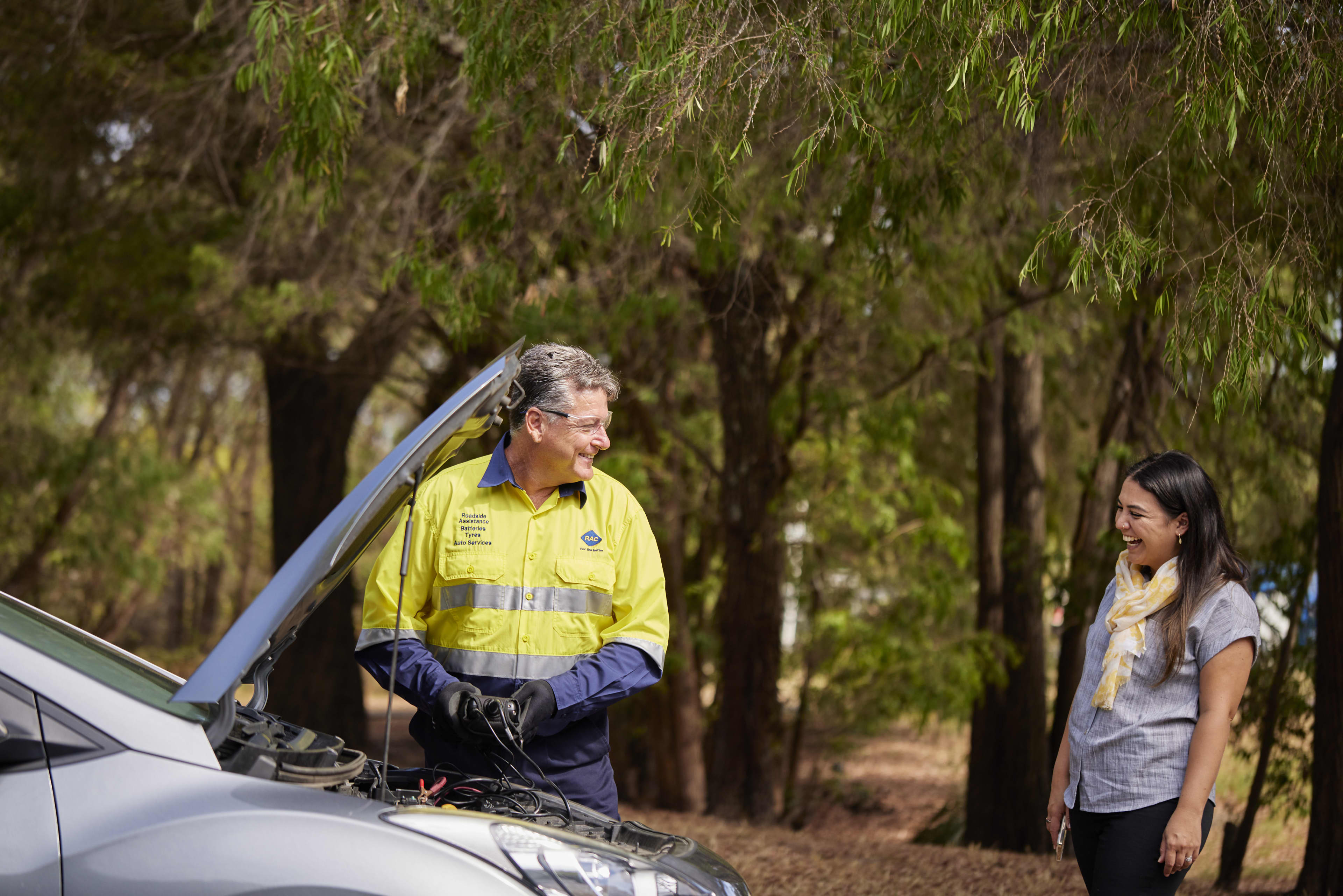 An older male roadside assistance technician wearing a high-visibility yellow and blue uniform, glasses, and black gloves smiles while working under the open hood of a silver car. A young woman with long dark hair, a grey shirt, and a yellow patterned scarf stands nearby, smiling back at him in a wooded, outdoor setting.