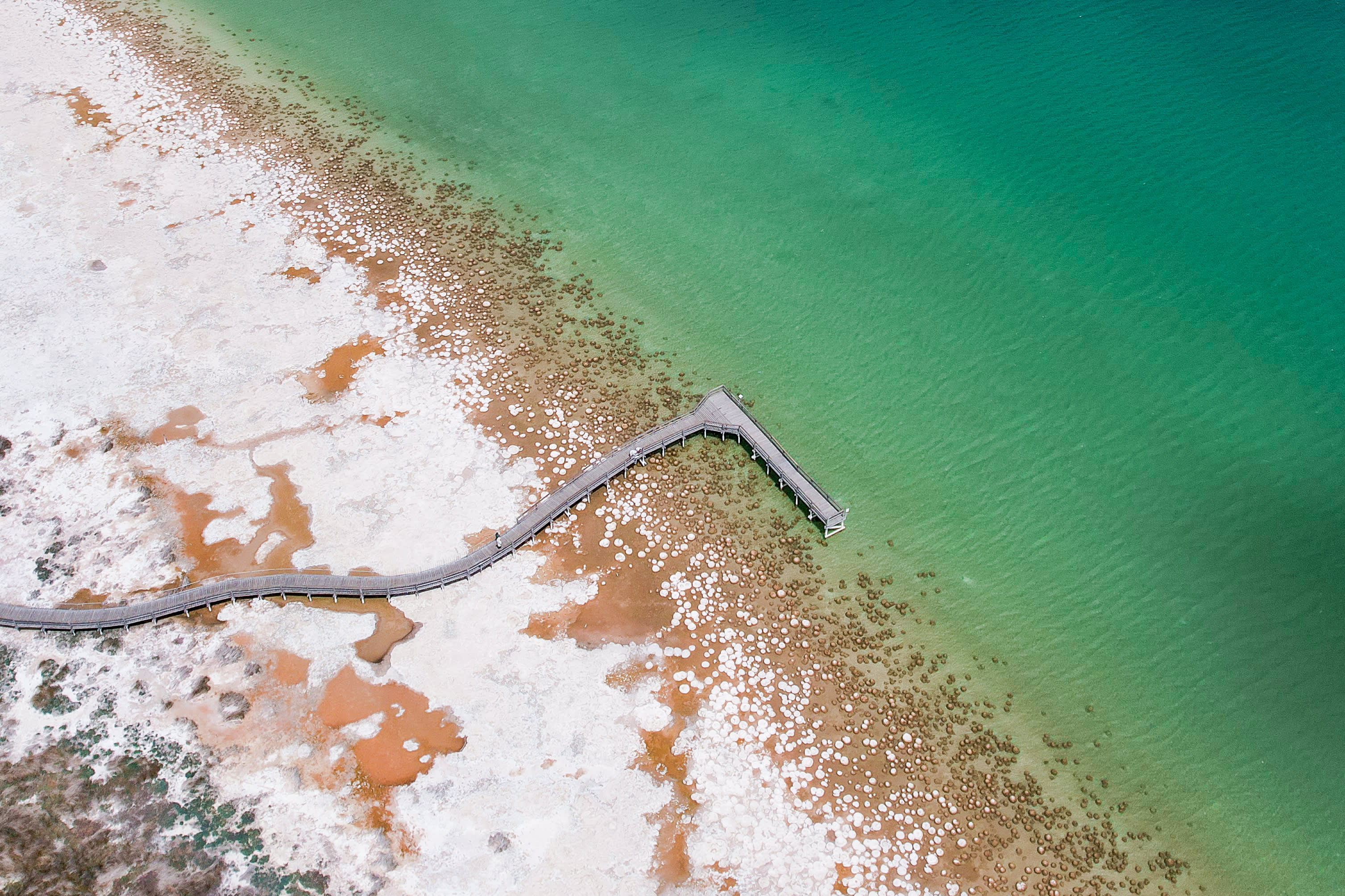 An aerial, top-down view shows a winding wooden boardwalk extending from a sandy, textured white and orange shoreline into clear, vibrant turquoise water. Scattered along the shallow edge of the water and across the shore are numerous rounded, rock-like thrombolite structures.