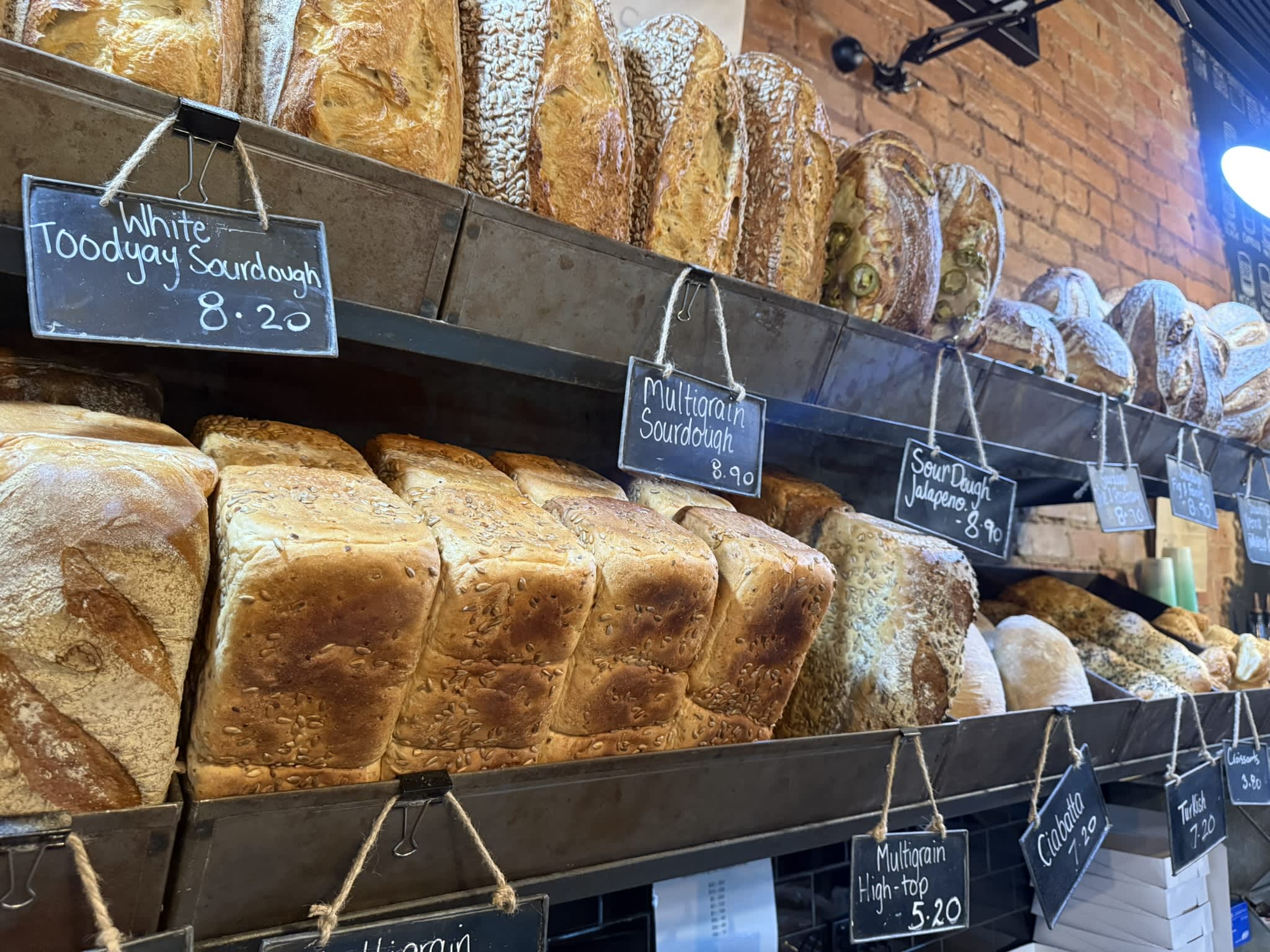 Loaves of artisanal bread displayed on rustic metal shelves in a bakery. The top shelf holds round and oblong loaves, including sourdough varieties. Small black chalkboard signs hang by twine in front of the bread, showing handwritten names and prices such as "White Toodyay Sourdough 8.20" and "Multigrain Sourdough 8.90." The middle shelf contains several rectangular, seed-crusted sandwich loaves arranged side by side. An exposed brick wall is visible in the background, lit by warm indoor lighting.