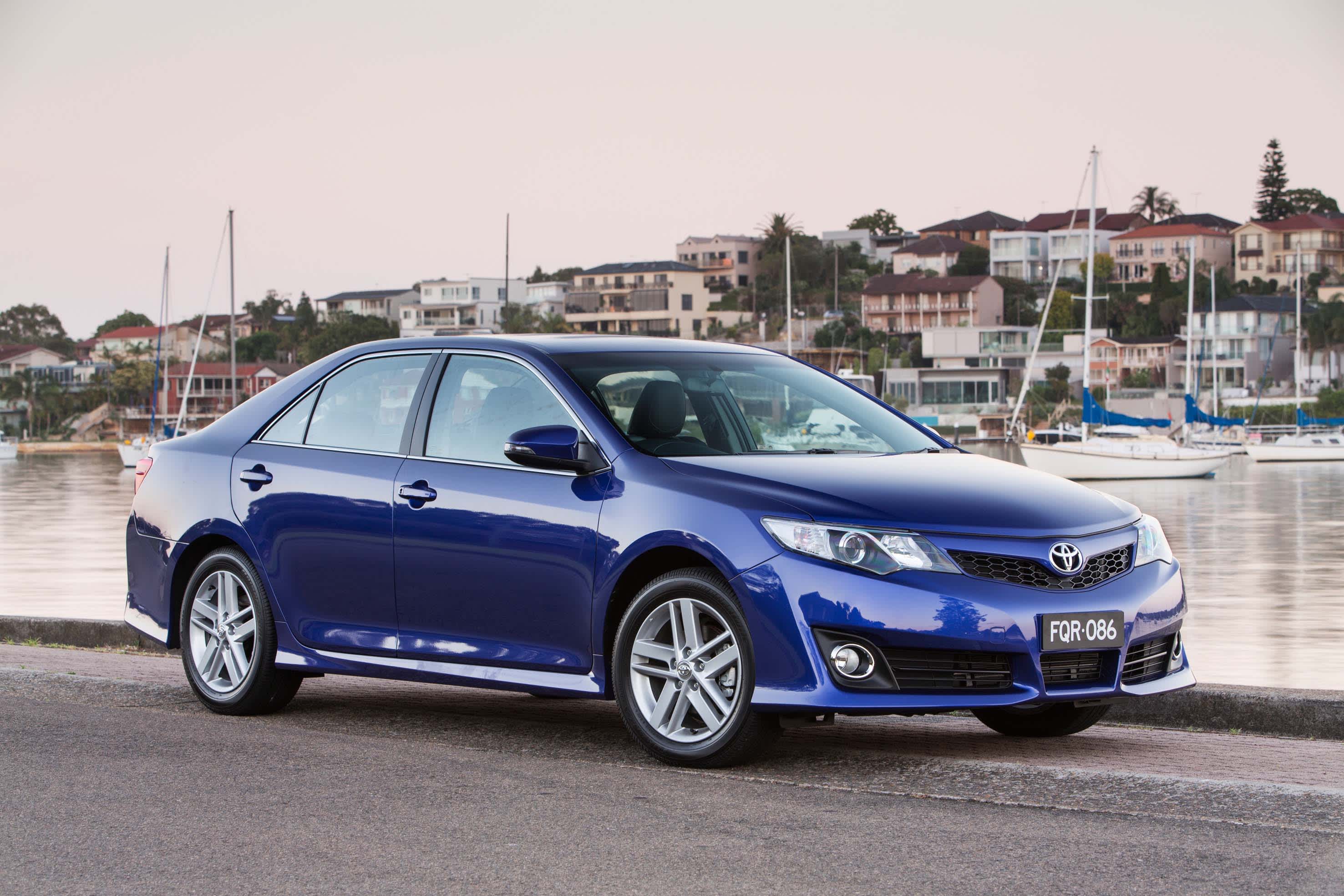 A dark blue Toyota Camry parked near a river with boats and homes in the background