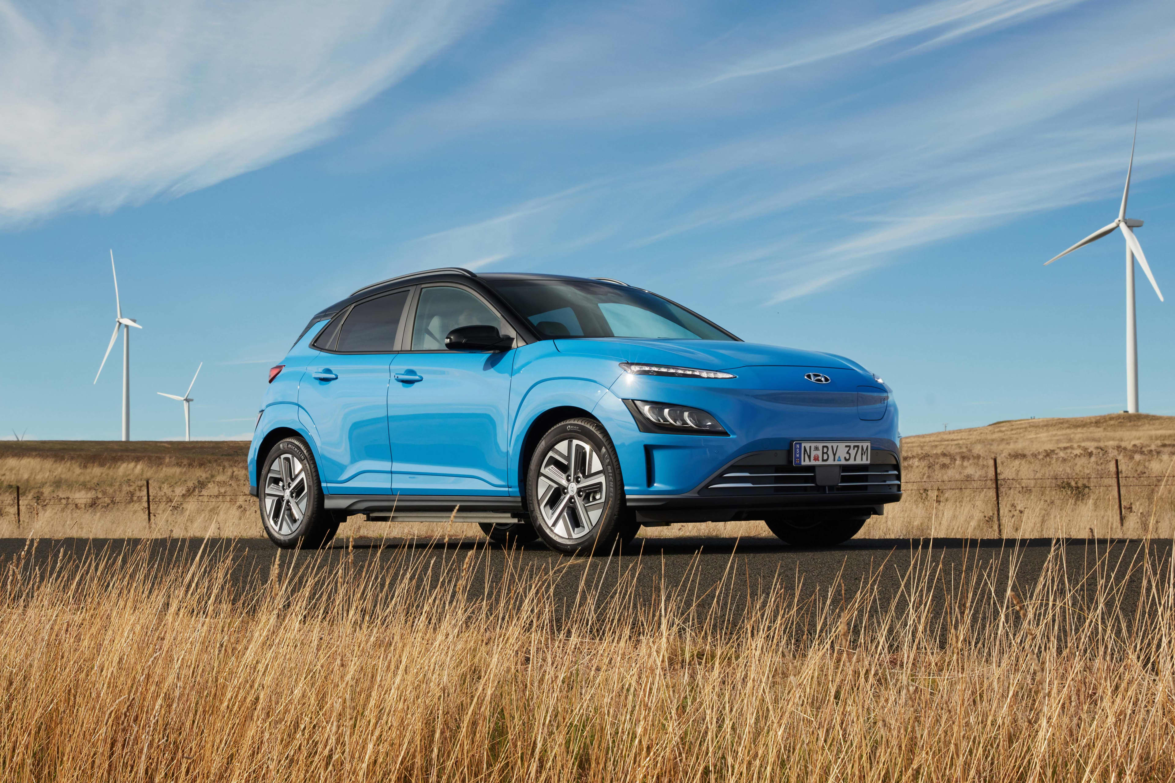 A blue Hyundai Kona EV parked in front of wind turbines.