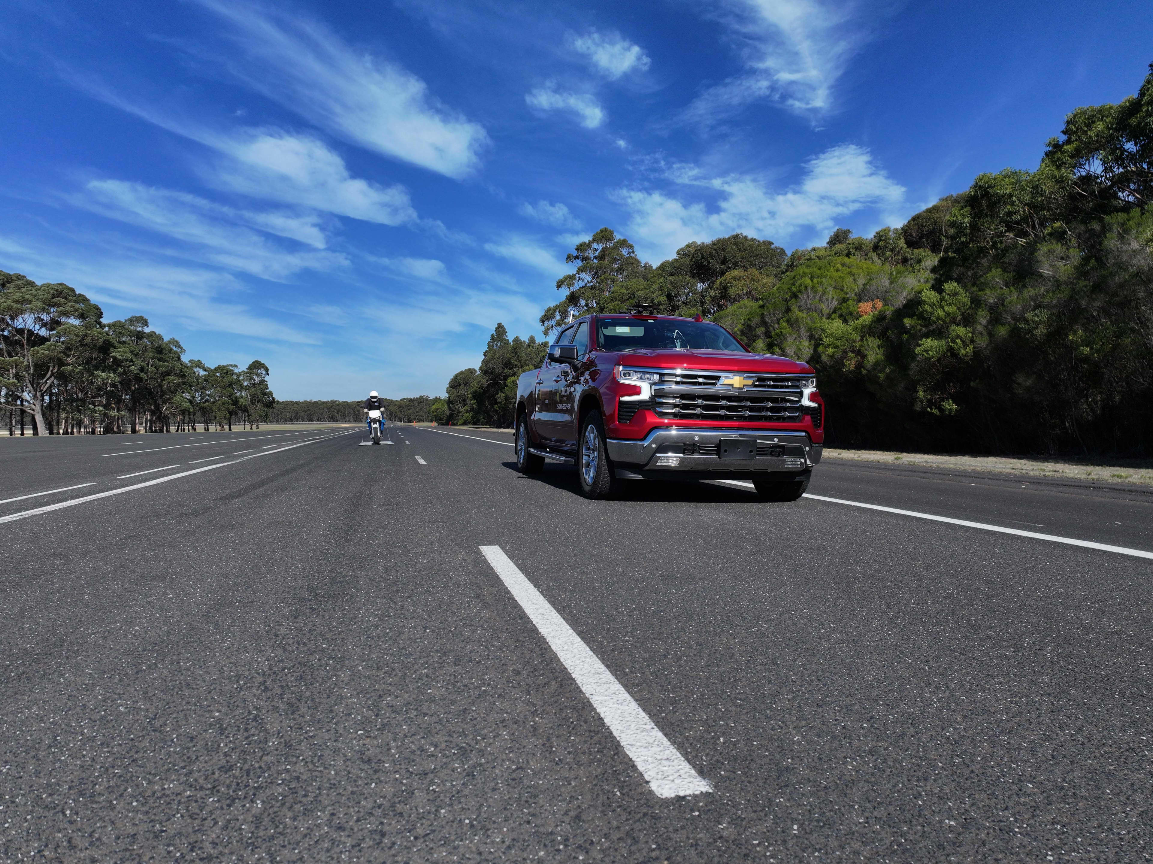 A red Chevrolet Silverado driving along a bitumenised country road with blue skies above.