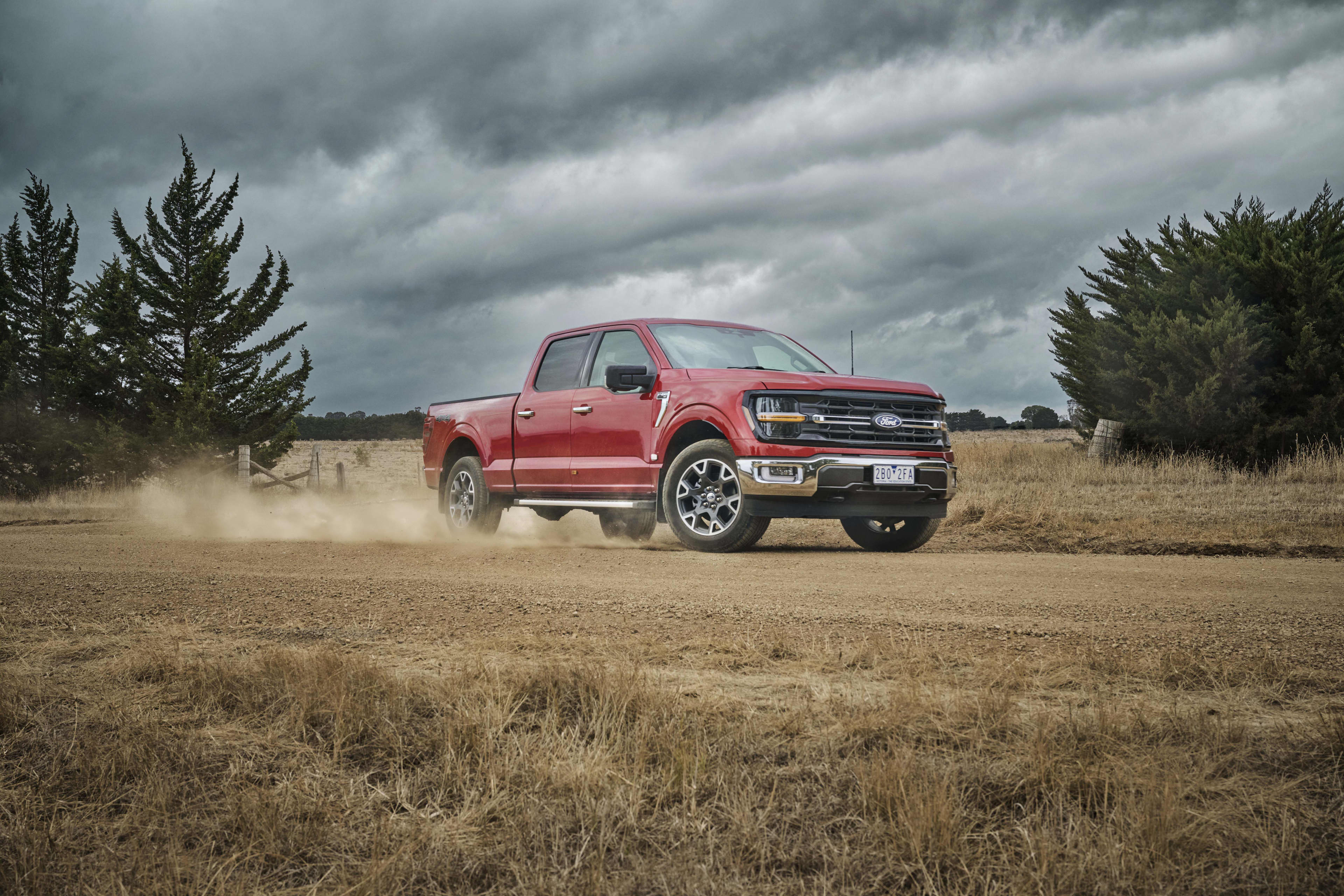A red Ford F-150 kicking up dust while driving along an unsealed country road.