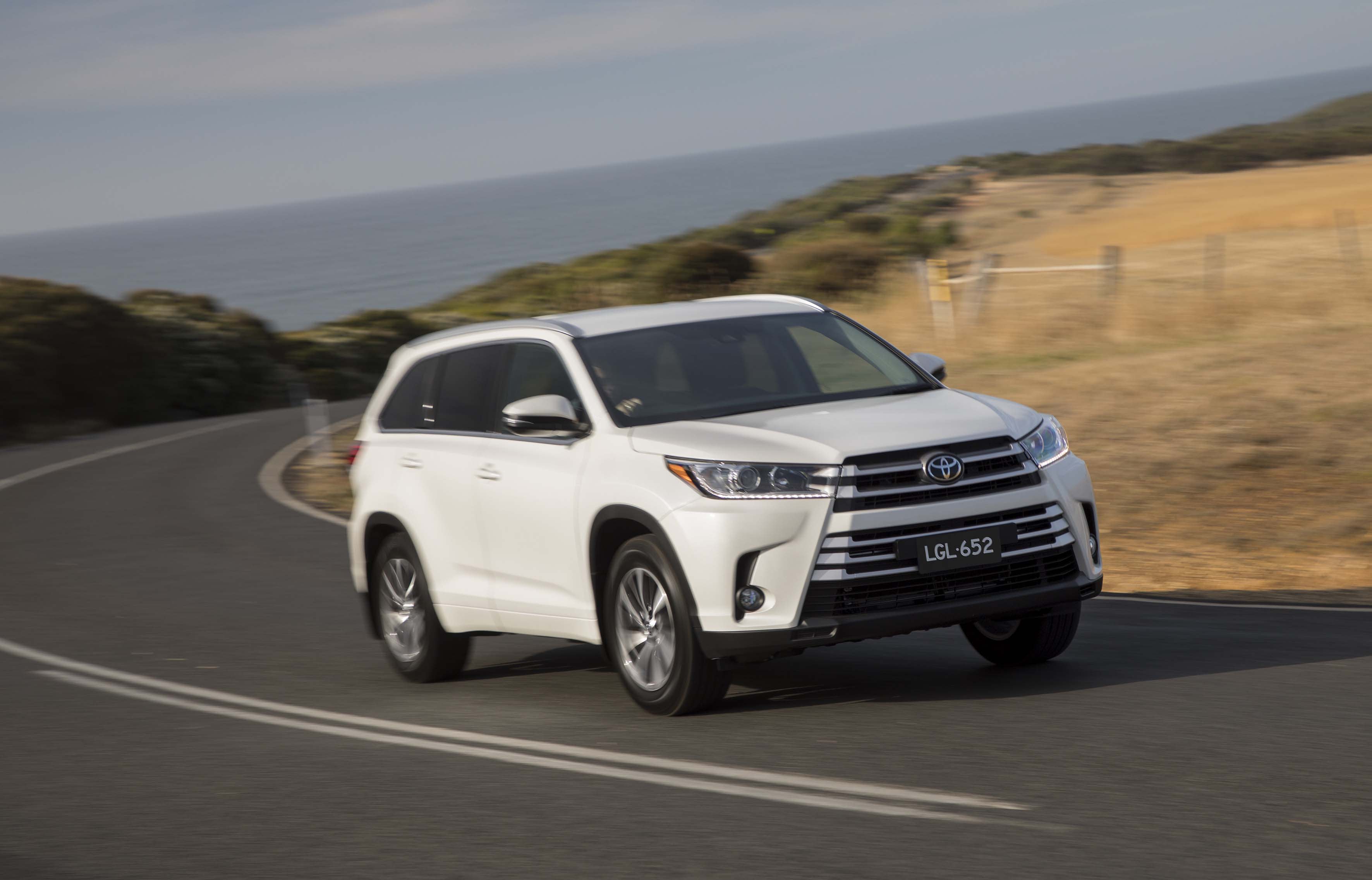 A white Toyota Kluger driving on a bend in a bitumen road in a country setting.