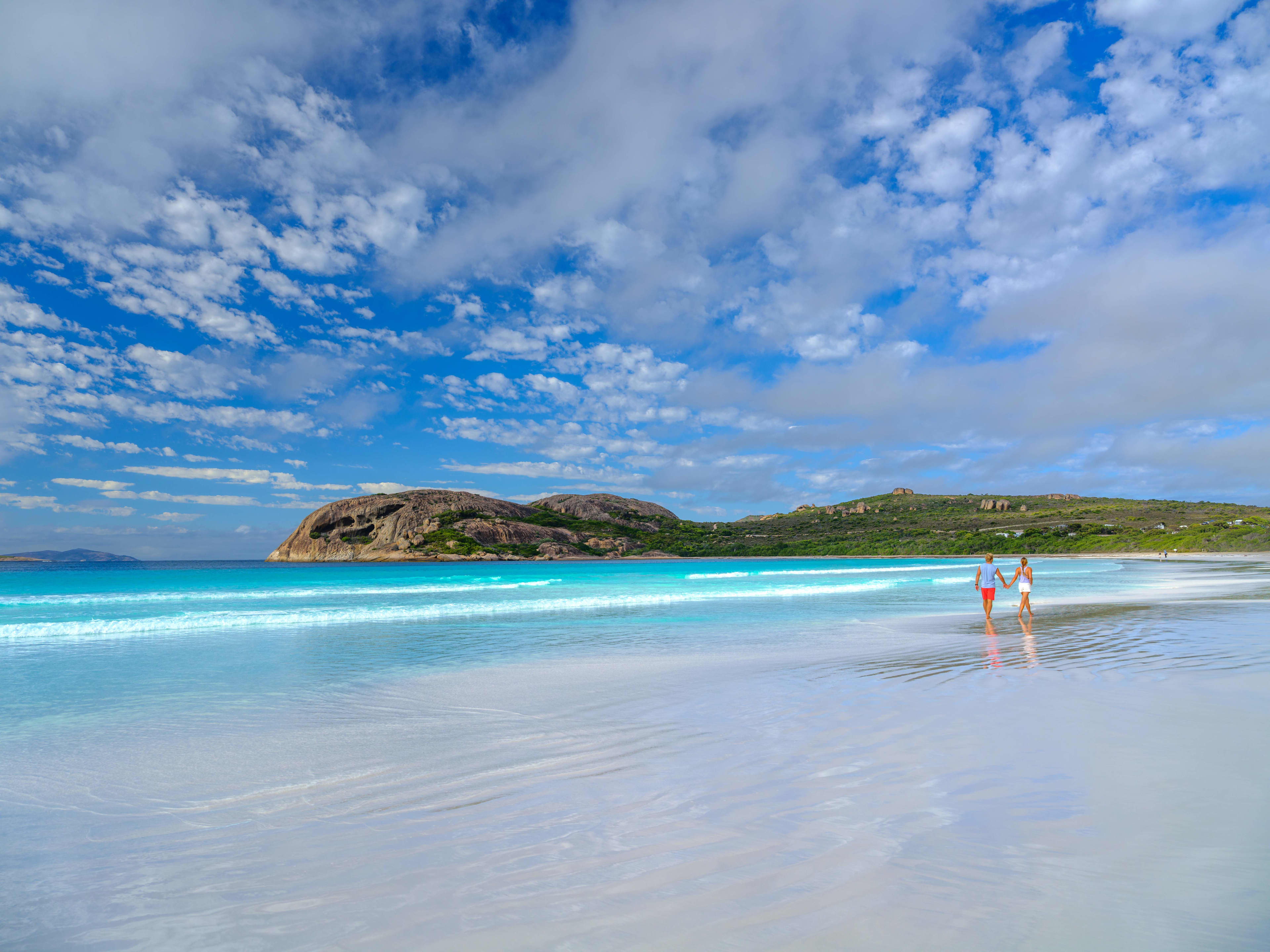 A couple hold hands and walk along the white sand at Lucky Bay with blue water and a cloudy blue sky.