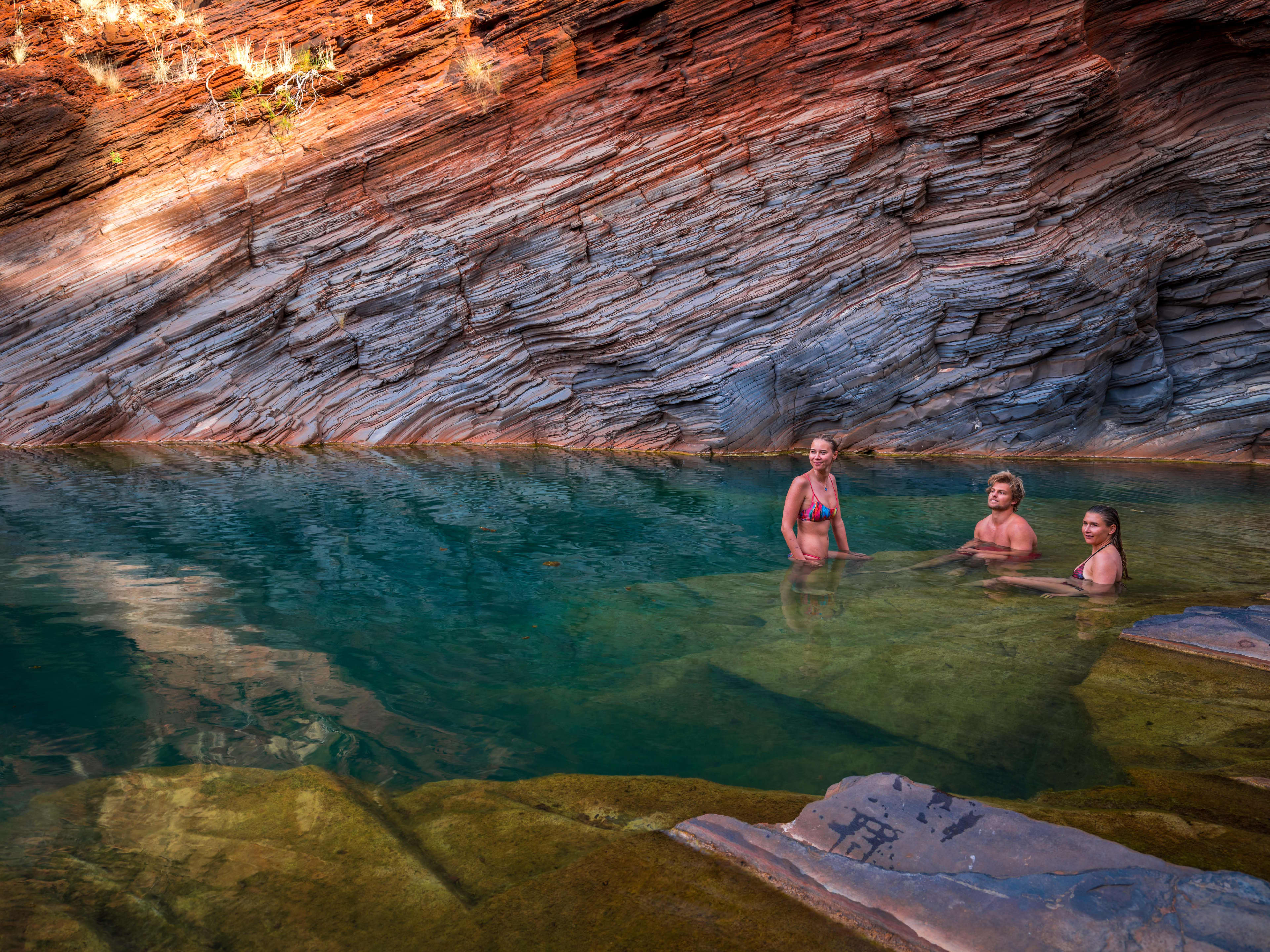 Two women and a man sitting in the blue water at the bottom of a rock gorge in Karijini National Park.