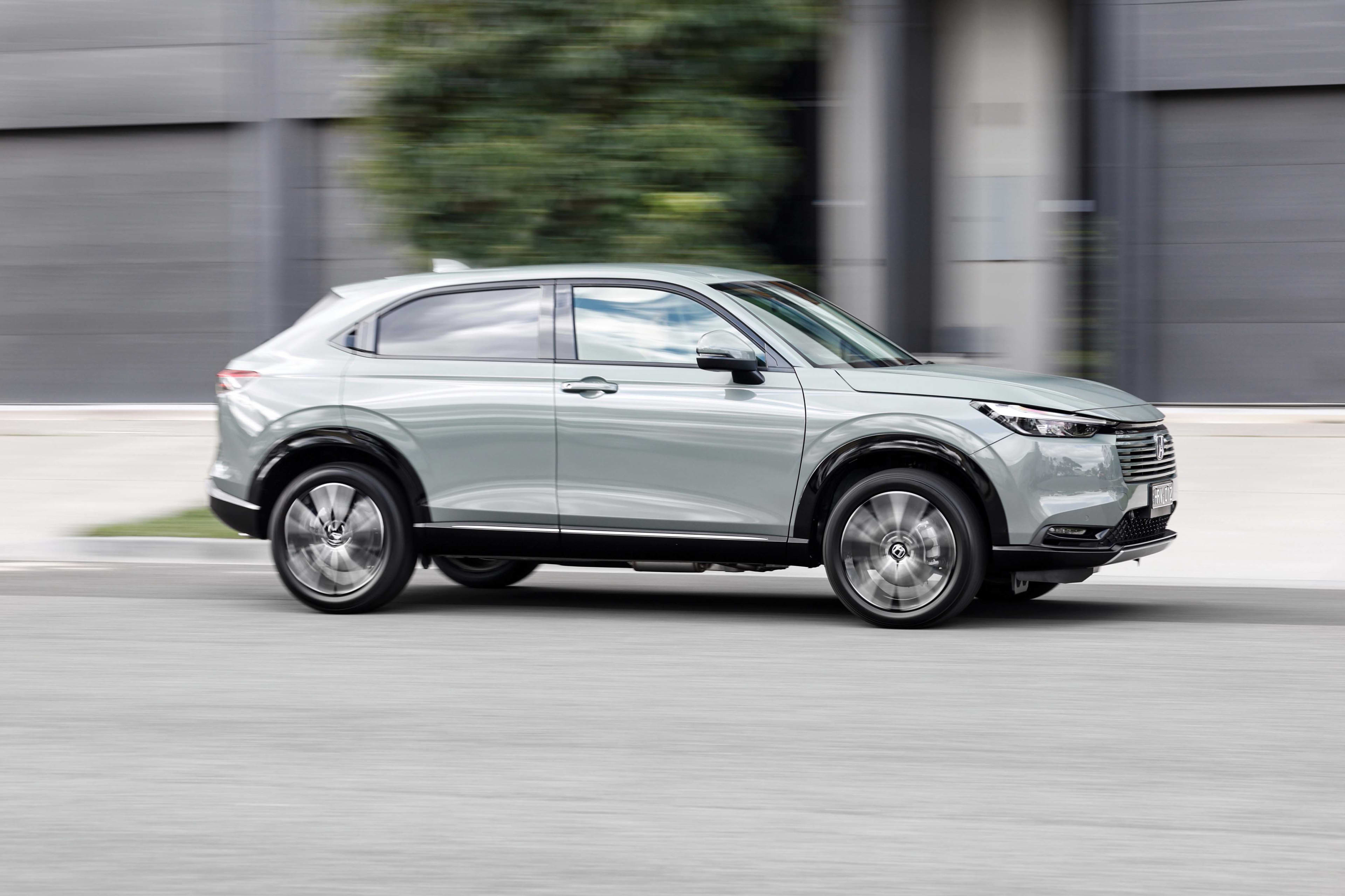 A silver Honda HR-V HEV driving along with blurred trees and building in the background.