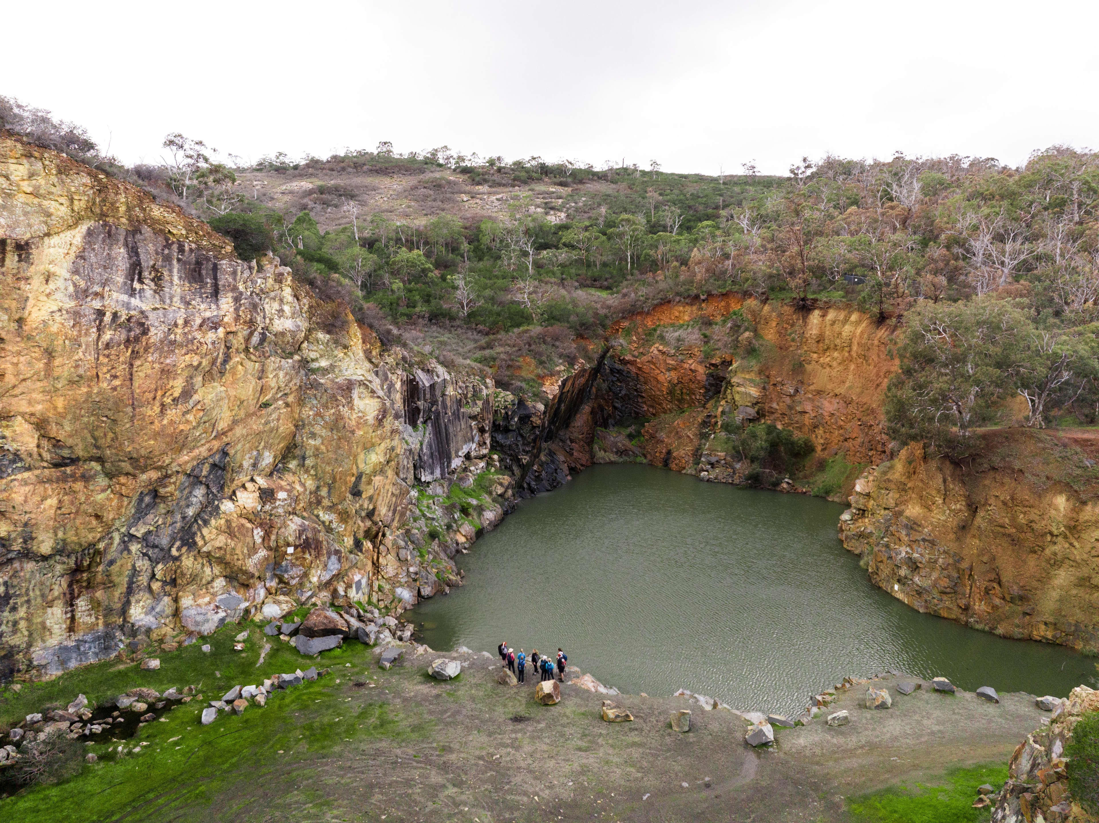 An elevated, wide shot shows a small group of people standing near a green-colored lake inside a steep rock quarry. The quarry features tall, rocky cliffs with yellow, orange, and grey rock faces. Green grass and scattered rocks cover the ground near the people. Trees and sparse vegetation grow along the upper edges of the quarry under an overcast sky.