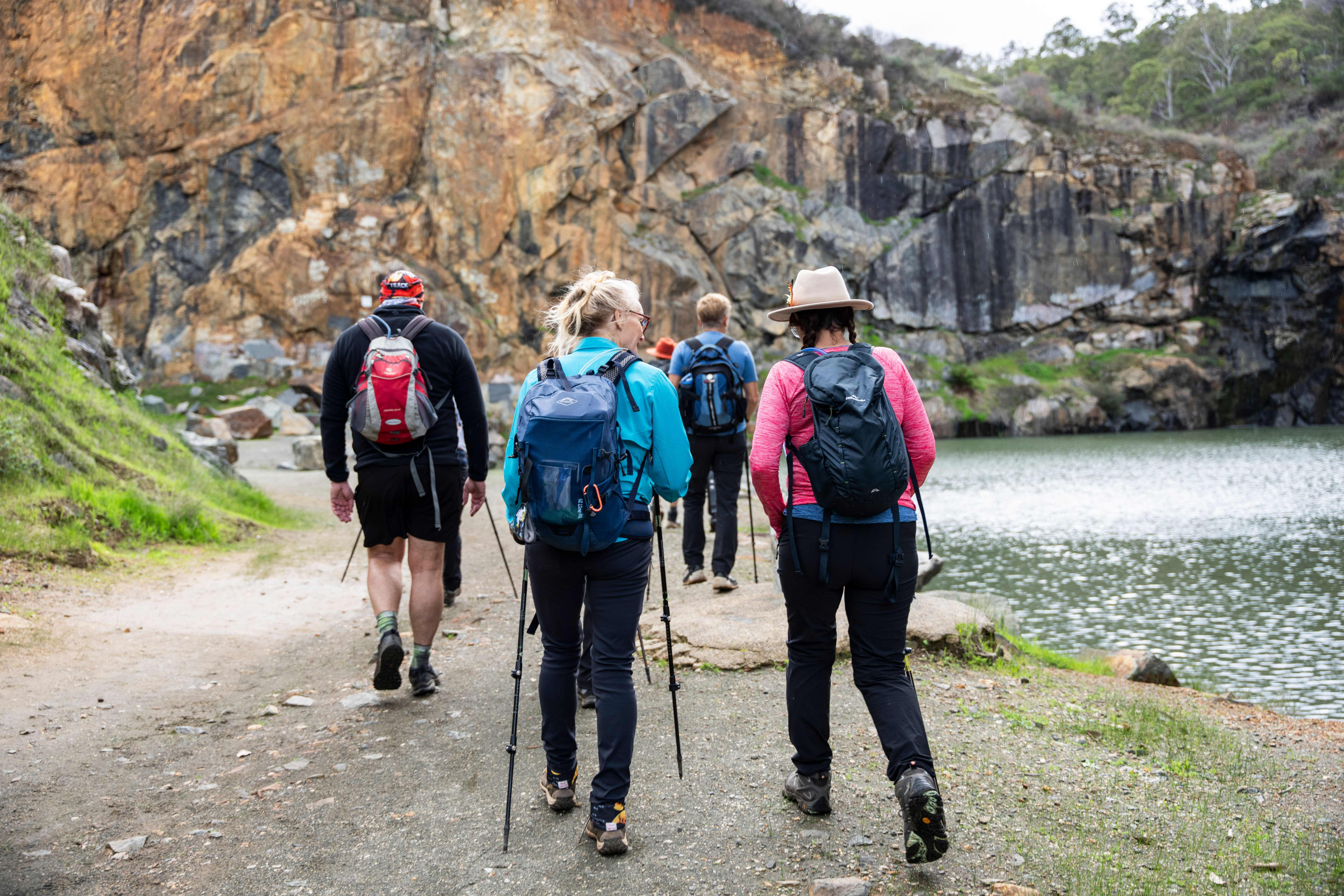 A group of hikers with backpacks walks along a dirt path next to a quarry lake, viewed from behind. The hikers are wearing athletic clothing, and some are using trekking poles. The path runs alongside water on the right, leading toward tall, rugged rock cliffs in the background with patches of green vegetation.