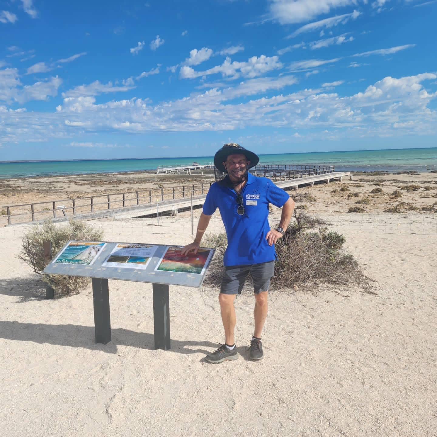 A campground host wearing a fly net over his face stands next to a sign on a beach.