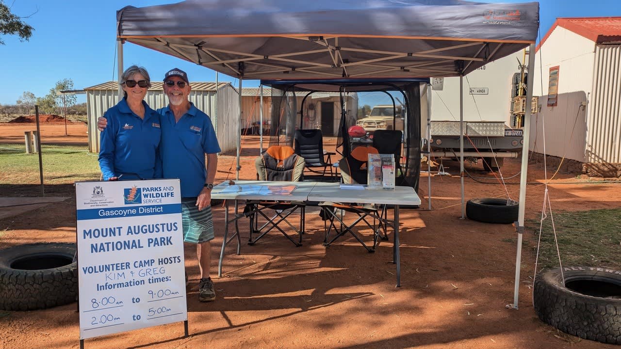 A couple of campground hosts, Kim and Greg, stand in front of an awning at a campground with a sign saying Mount Augustus National Park.
