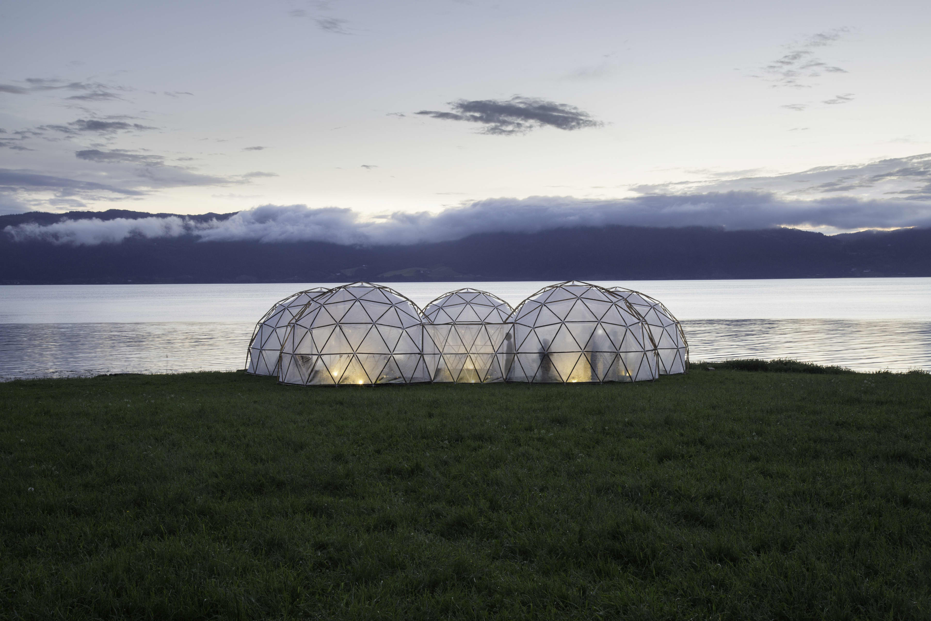 Geodesic dome structures glow at dusk on a grassy lakeshore, with calm water, mist-covered mountains, and a cloudy sky in the background.
