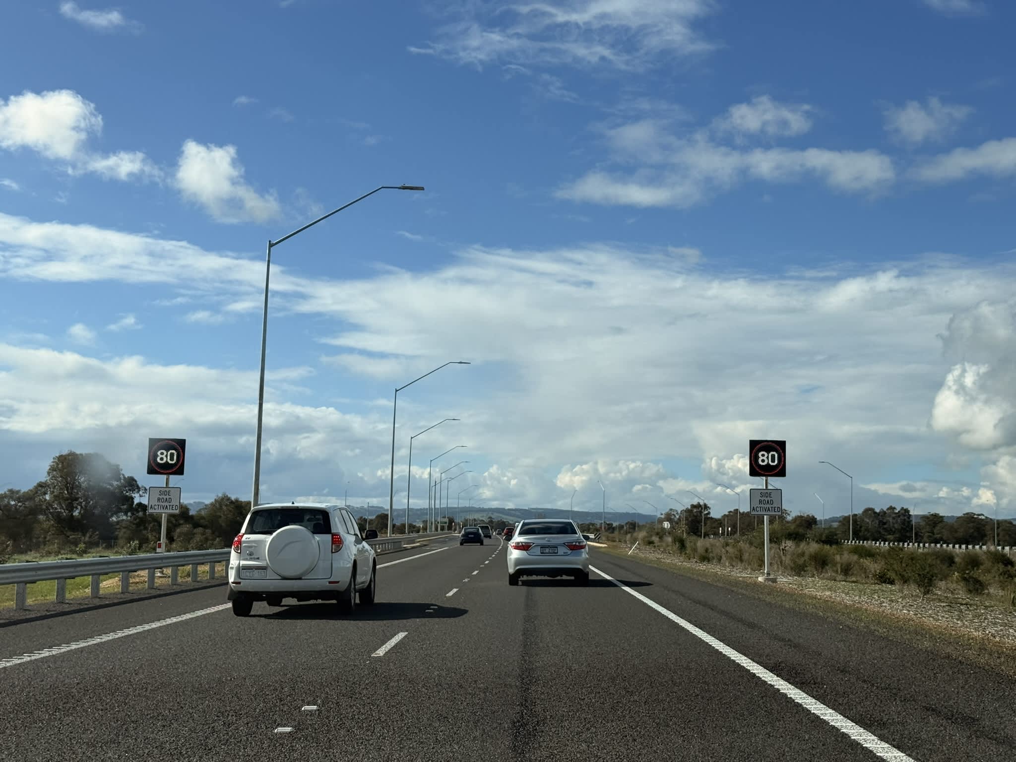 Two cars on a WA highway pass electronic signs showing the speed 80kmh with the words 'side road activated' on a placard underneath.