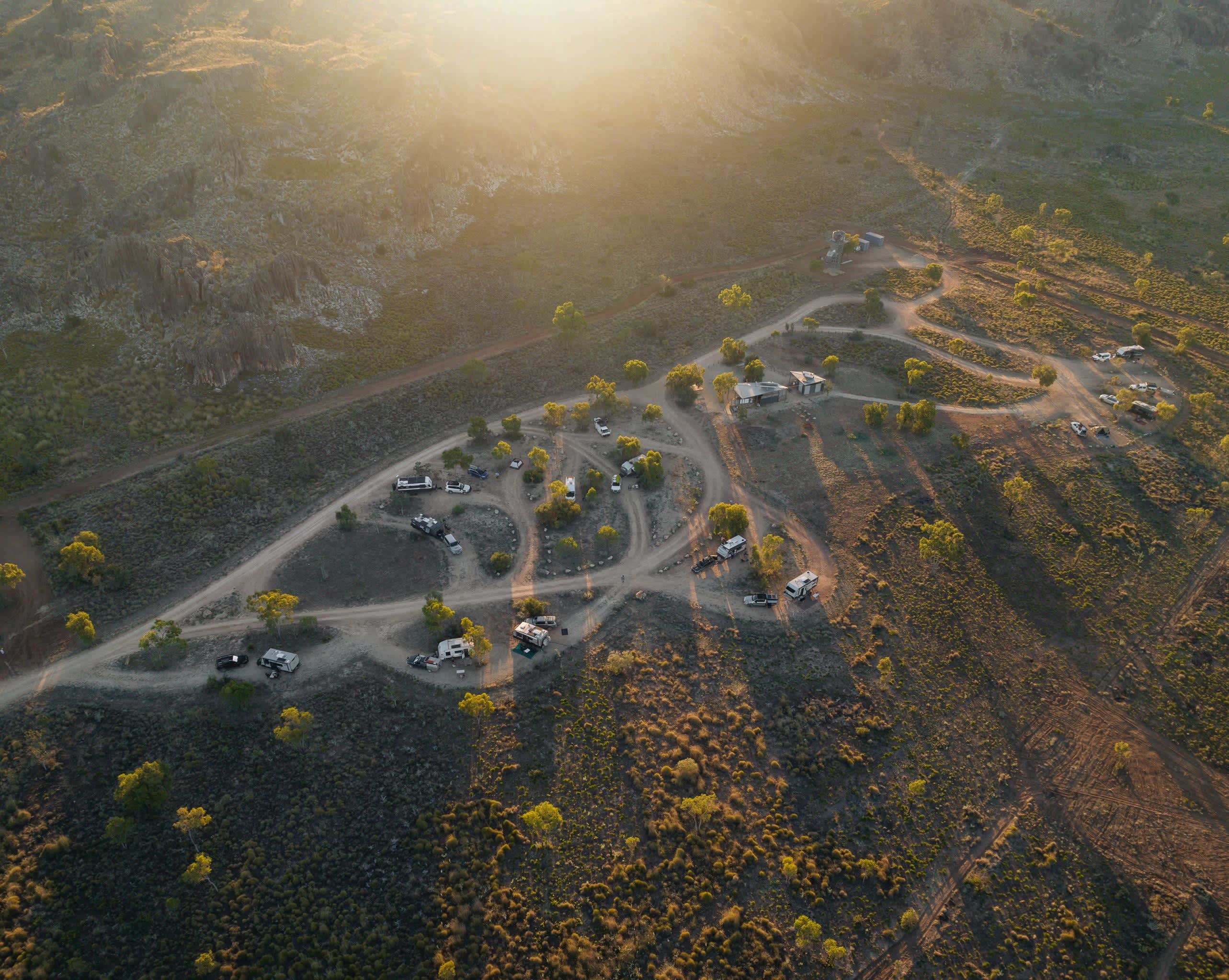 An aerial, high-angle view captures a campgrounds loop nestled in a vast, arid landscape bathed in the warm, golden light of either sunrise or sunset. The campground features a network of dusty, winding dirt roads that form a distinct tear-drop or leaf shape, with individual campsites carved out along the loops. Several camper vans, RVs, and trucks are parked at various sites, interspersed with sparse green trees and low-lying desert shrubs. In the background, rugged, rocky hills rise up, with the bright sun flaring over the ridge, casting long, soft shadows across the entire terrain.