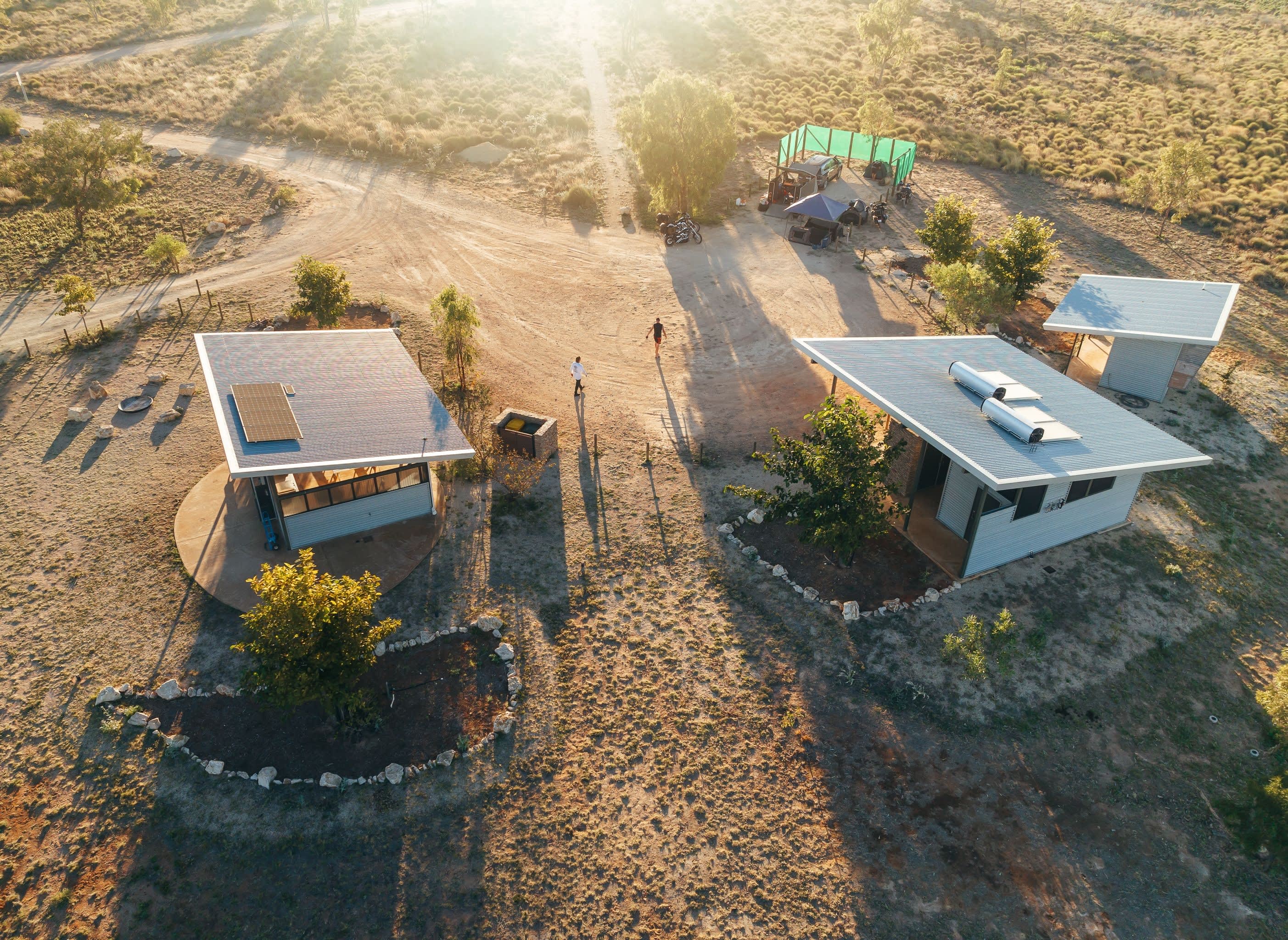 An aerial, high-angle view shows modern eco-cabins with slanted corrugated metal roofs in an arid, sun-drenched landscape. The structures feature solar panels and water heating systems, casting long shadows across the dry, sandy ground dotted with sparse patches of grass and small green trees. In the background, two people are walking along a dirt pathway toward a camping setup with a green canopy shield, all illuminated by the bright, hazy glow of a low sun.