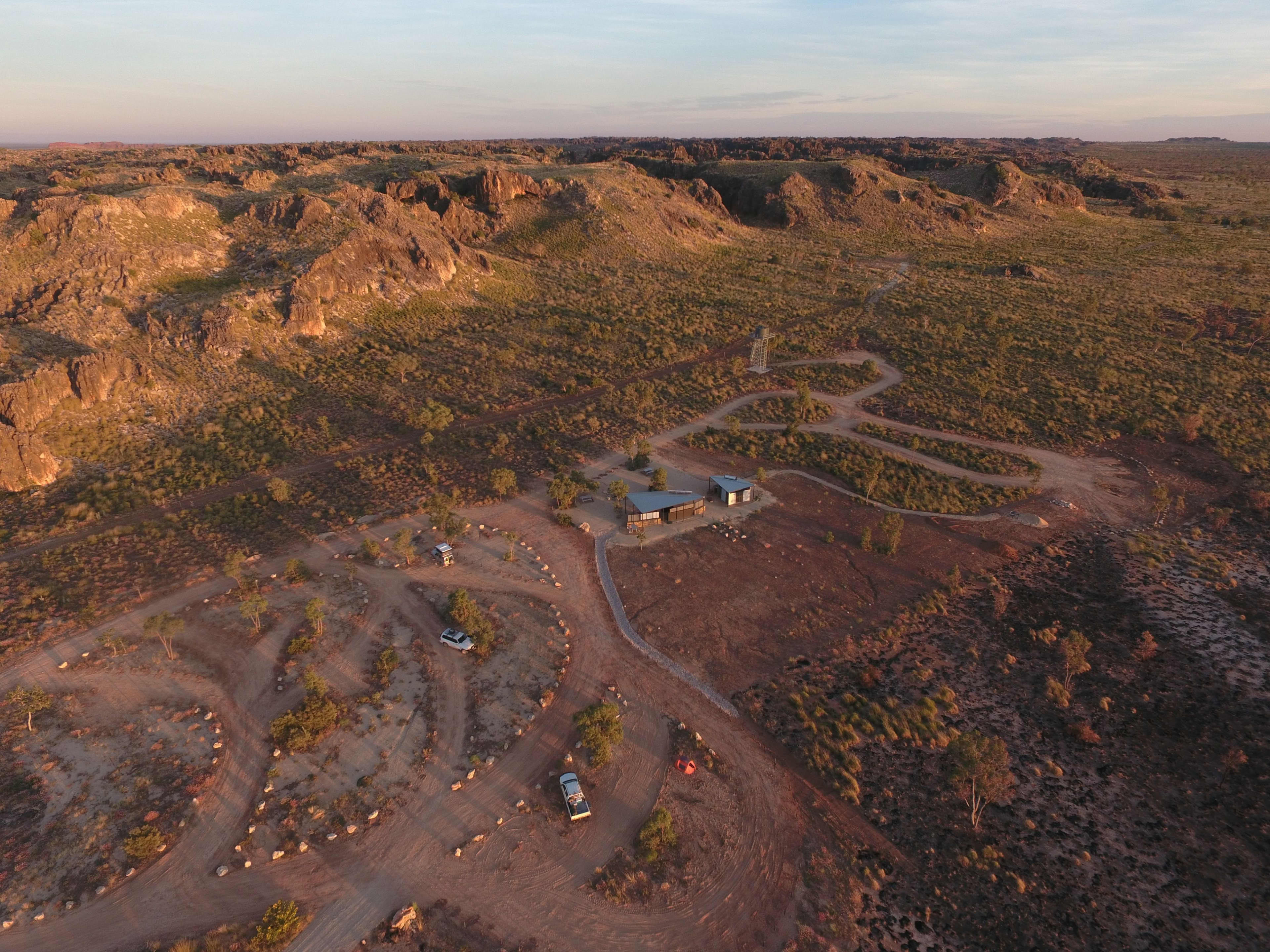 An aerial, high-angle view reveals a remote campground and facility nestled at the base of vast, rugged rocky hills. The landscape is dominated by dry, reddish-brown earth and sparse green vegetation, all softly illuminated by the warm light of early morning or late afternoon. Dirt tracks and a paved pathway connect a cluster of modern buildings to smaller, winding parking loops where a couple of vehicles are parked. The rocky ridges rise steeply in the background, stretching across the horizon under a clear, pale sky.
