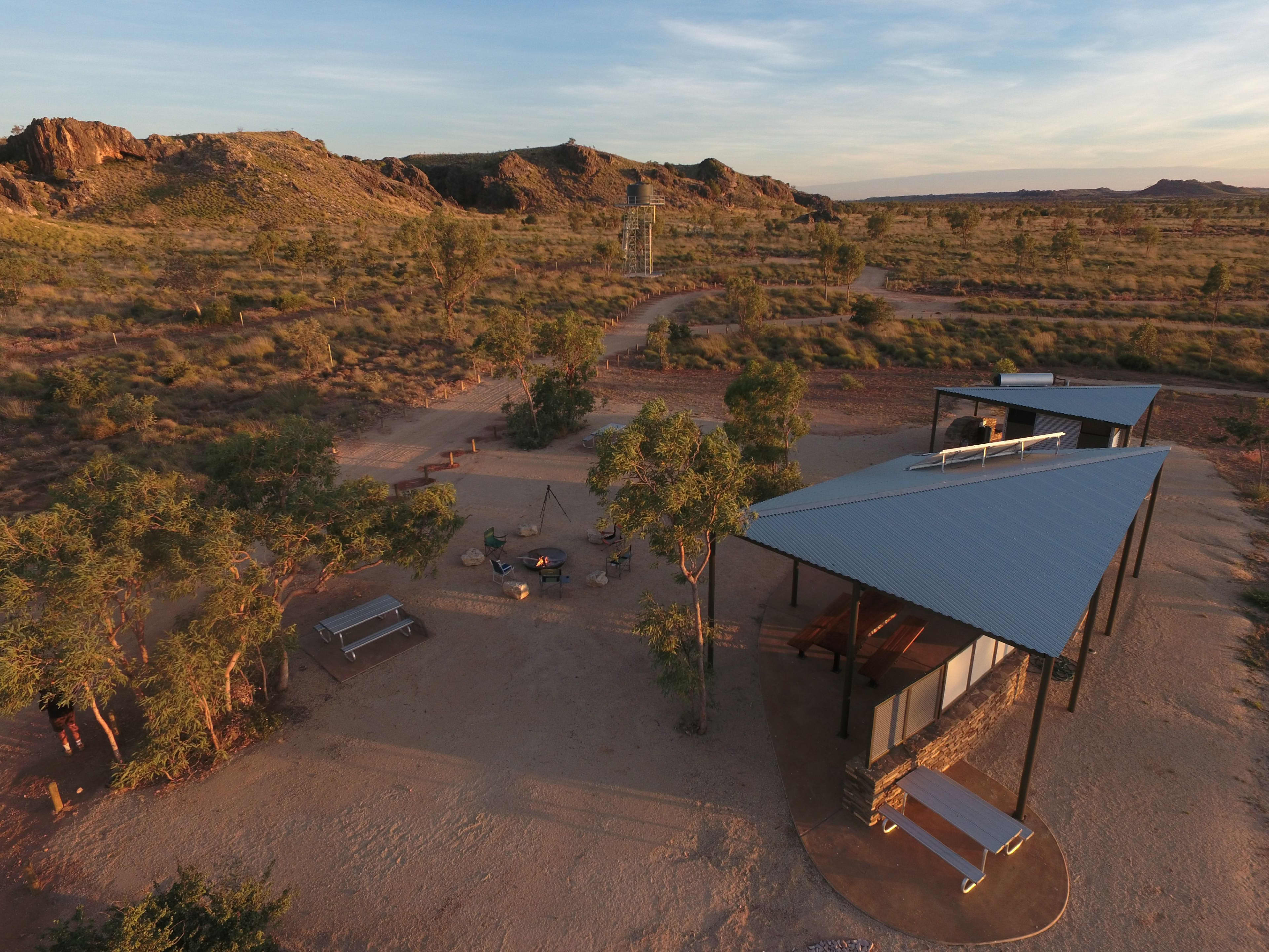 An aerial, high-angle view shows an outdoor communal area at a remote campground surrounded by a vast, rugged outback landscape. In the foreground, a modern open-air shelter features a slanted corrugated metal roof, a stone accent wall, and a metal picnic table. Nearby, a cleared dirt clearing includes a central campfire ring surrounded by portable lawn chairs, picnic tables, and sparse green trees. In the background, a walking path winds past a distant metal observation tower toward low, rocky hills under a wide sky with light, streaky clouds in the warm glow of late afternoon.