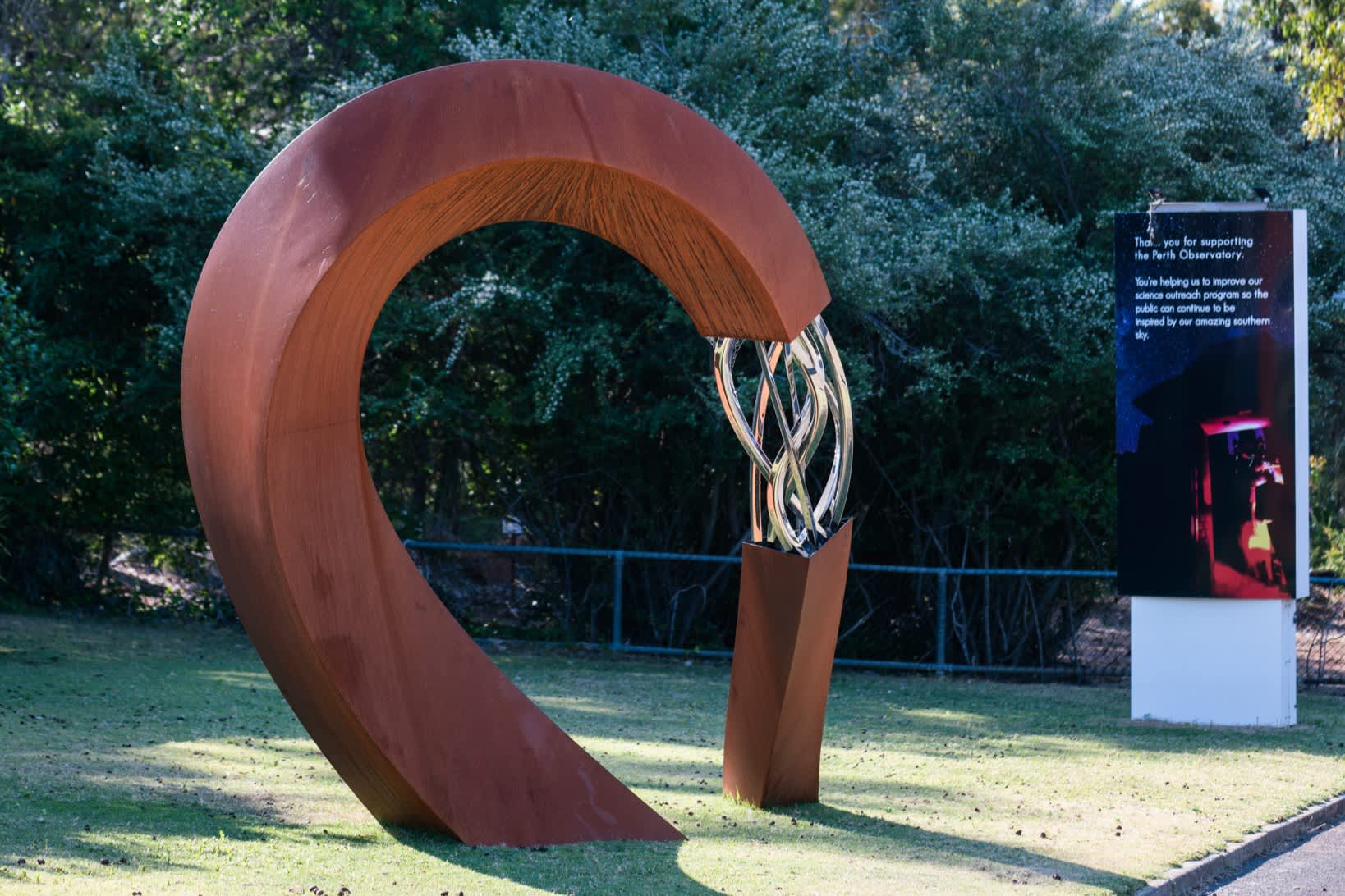 A large, modern C-shaped sculpture made of rusted weathering steel stands on a grassy lawn, with shiny, interwoven stainless steel ribbons filling the gap in its loop. In the background, green trees and bushes are visible behind a low fence, and a tall informational sign stands to the right.