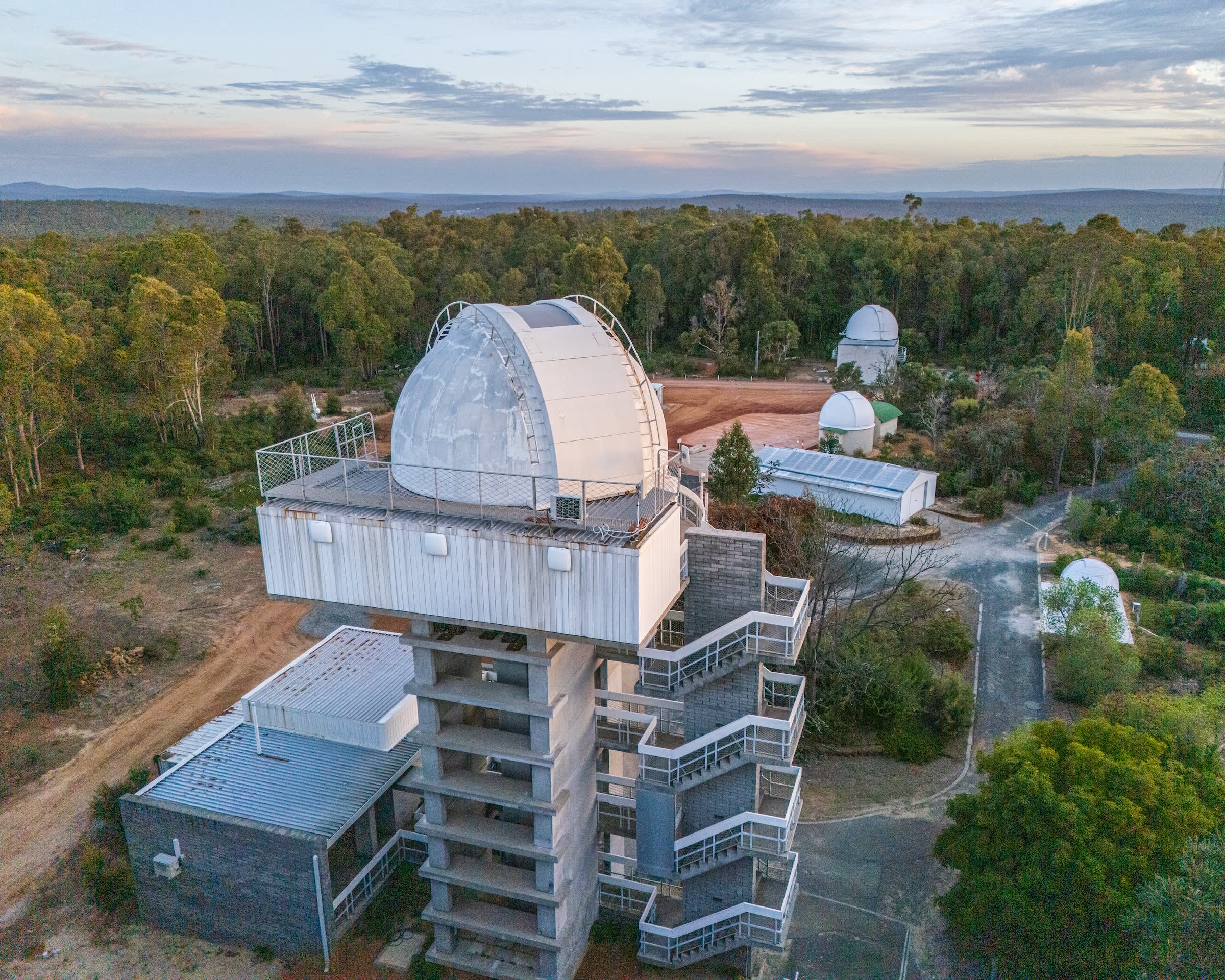 An aerial view shows a white, dome-topped observatory tower surrounded by a dense green forest under a cloudy, dawn sky. The tall, concrete structure in the foreground features an external zigzagging staircase leading to a high platform, while several smaller observatory domes and support buildings are scattered throughout the wooded hills in the background.