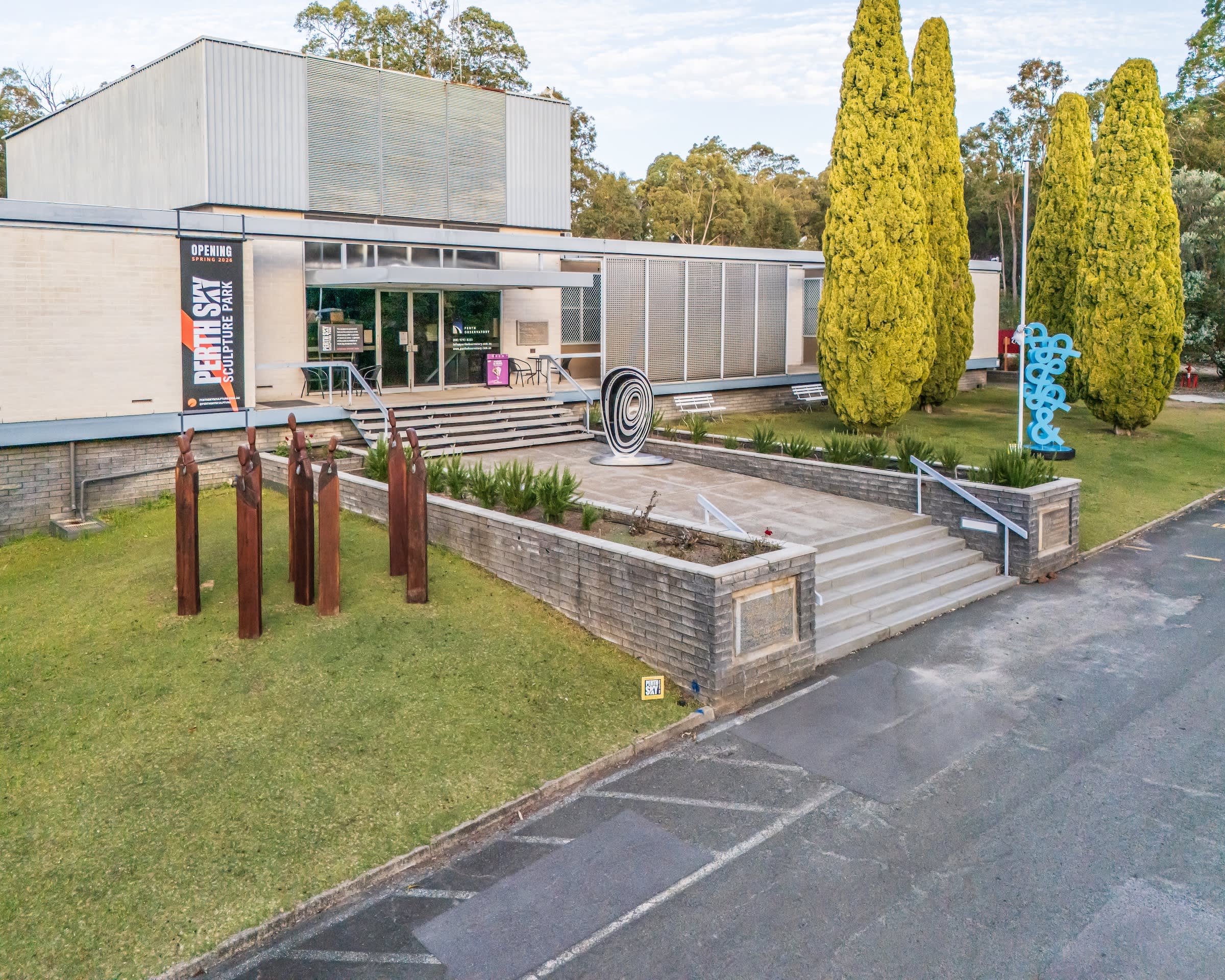 An exterior aerial view shows a modern single-story building labeled "Perth Sky Sculpture Park" with several outdoor art installations displayed on its grounds. To the left, a cluster of thin, rusted vertical metal posts stands on the lawn, while a black-and-white swirling spiral sculpture rests near the main stone entrance steps. On the right lawn, a tall teal-blue abstract looping sculpture stands next to manicured cypress trees against a backdrop of native forest.