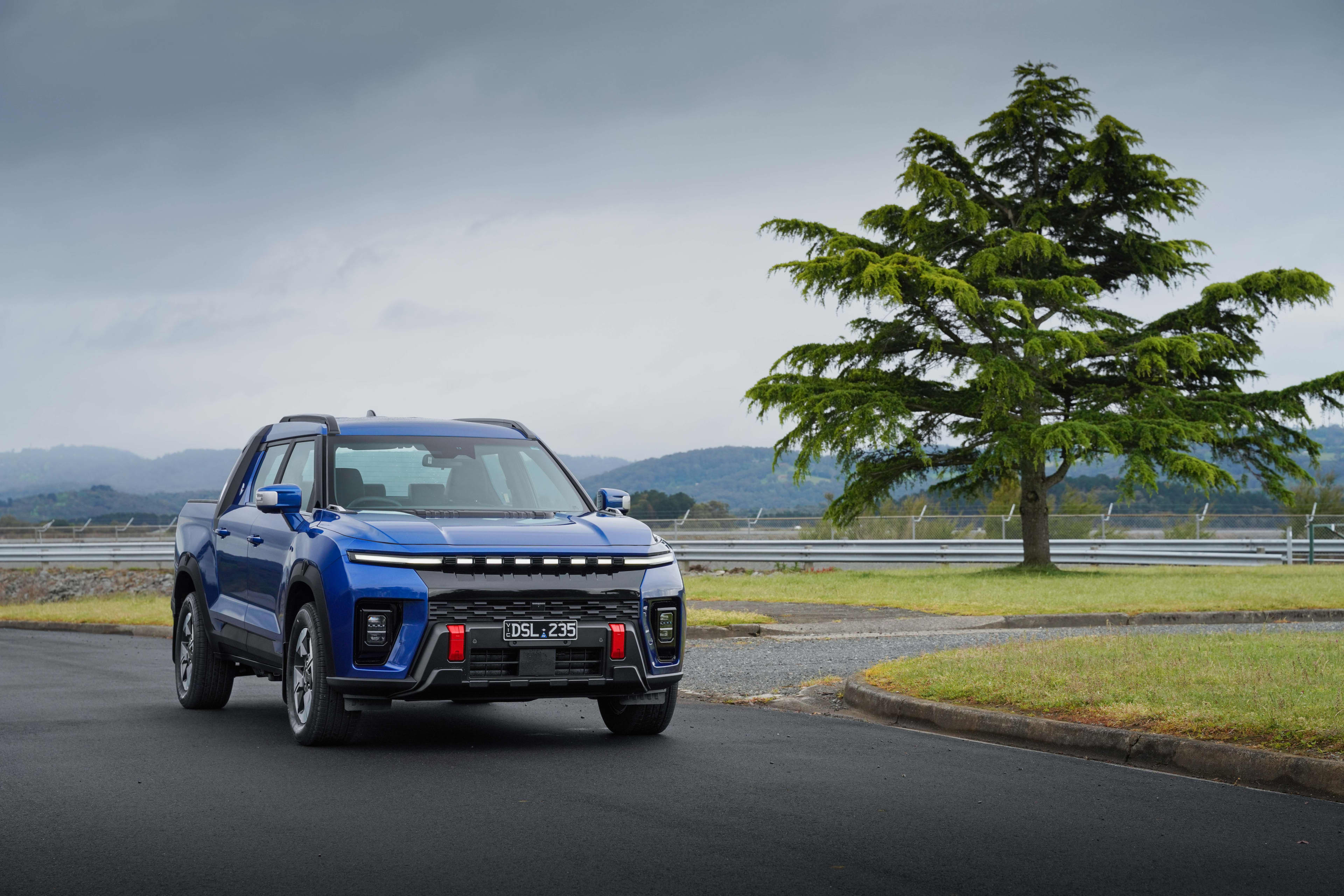 A blue KGM Musso EV ute driving on a country road with a tall tree in the background.