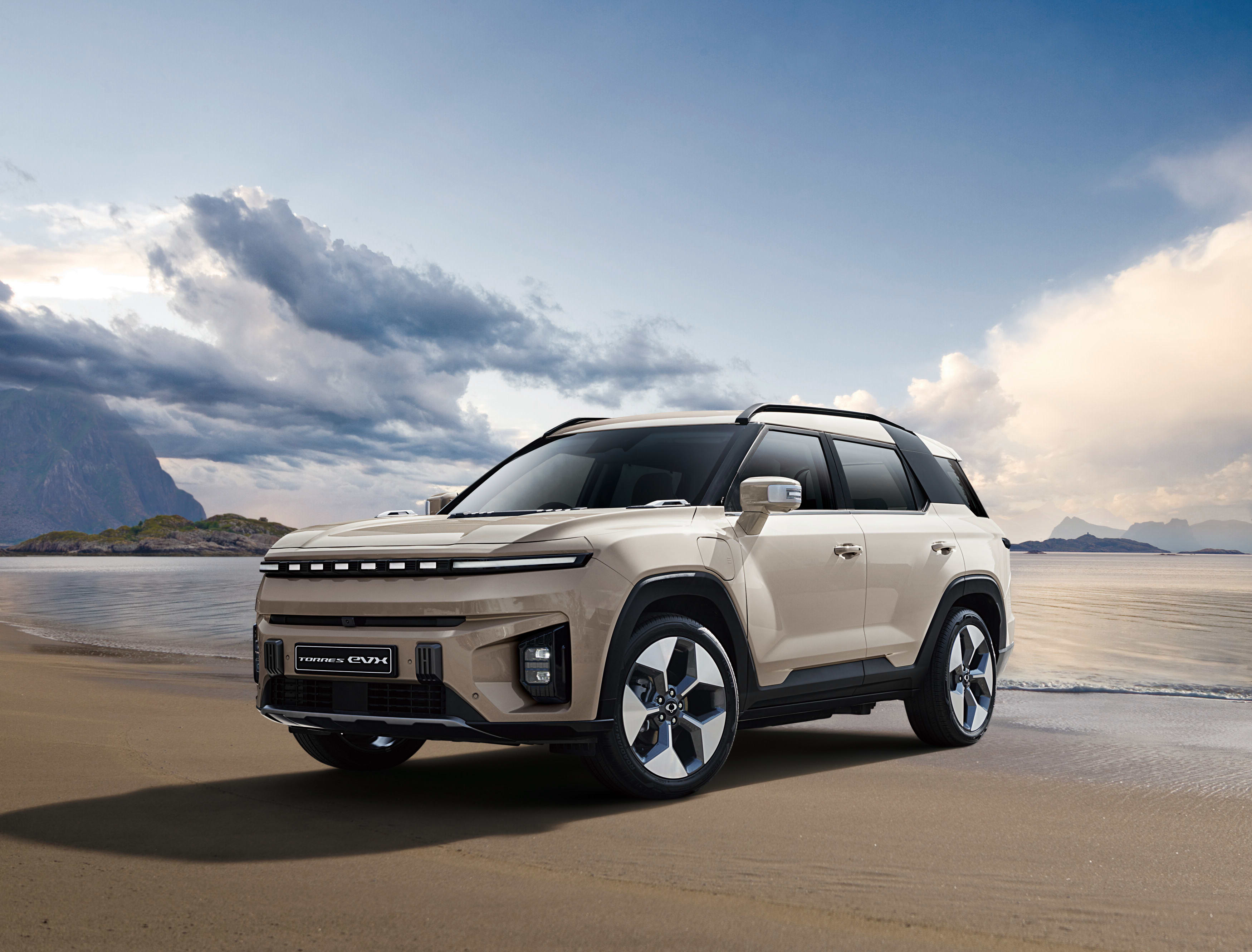 A white KGM Torres EVX parked on a beach with a cloudy sky in the background.