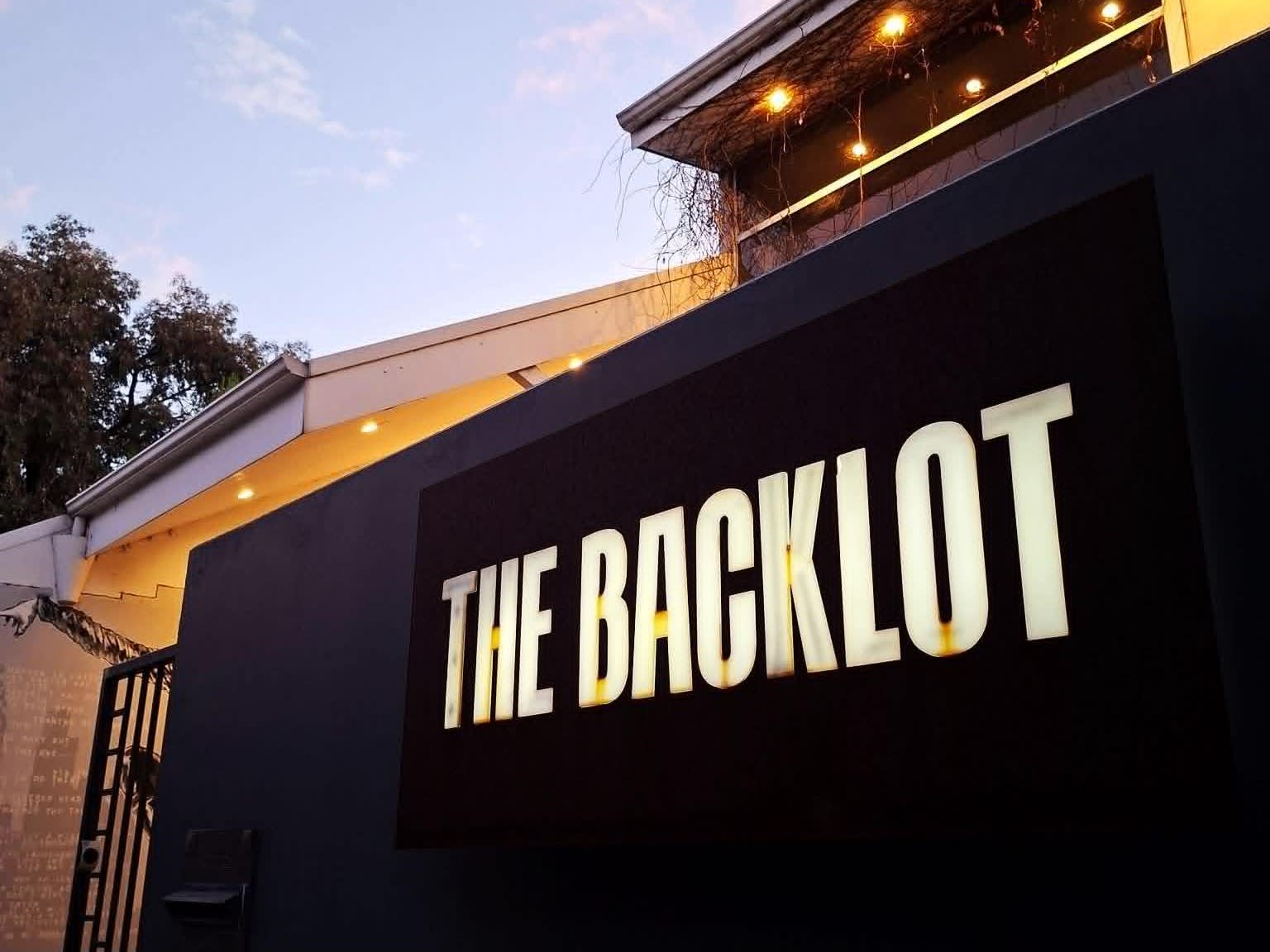 A glowing, rectangular sign reads "THE BACKLOT" in bold, capitalized white letters against a black background.