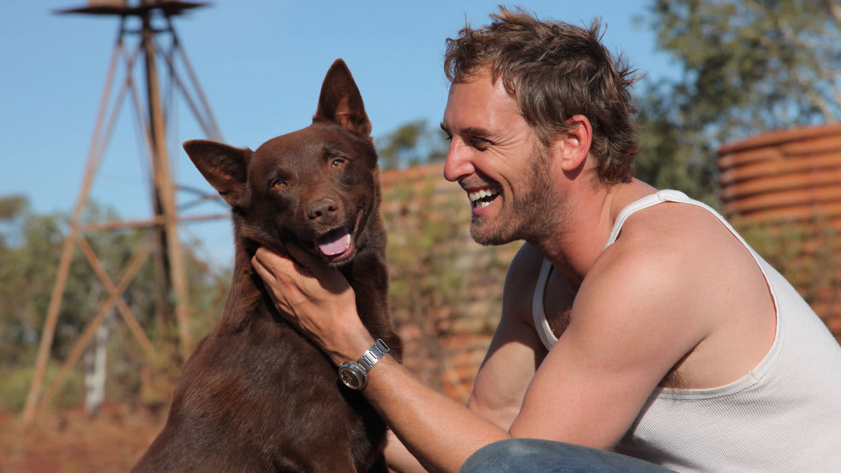 A smiling, muscular man wearing a white tank top crouches next to a chocolate-brown Australian kelpie in an outback setting.