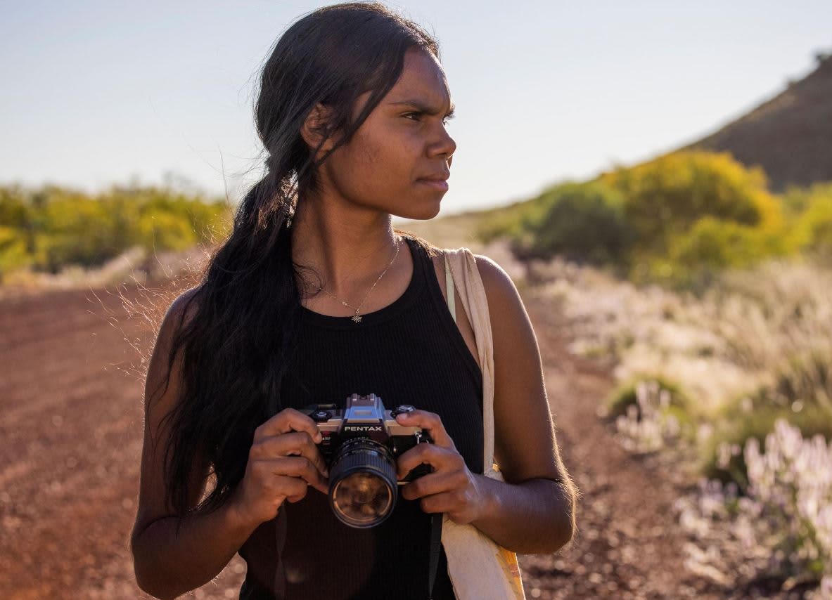 A side-profile view of a young woman with long, dark hair holding a vintage SLR film camera in her hands.