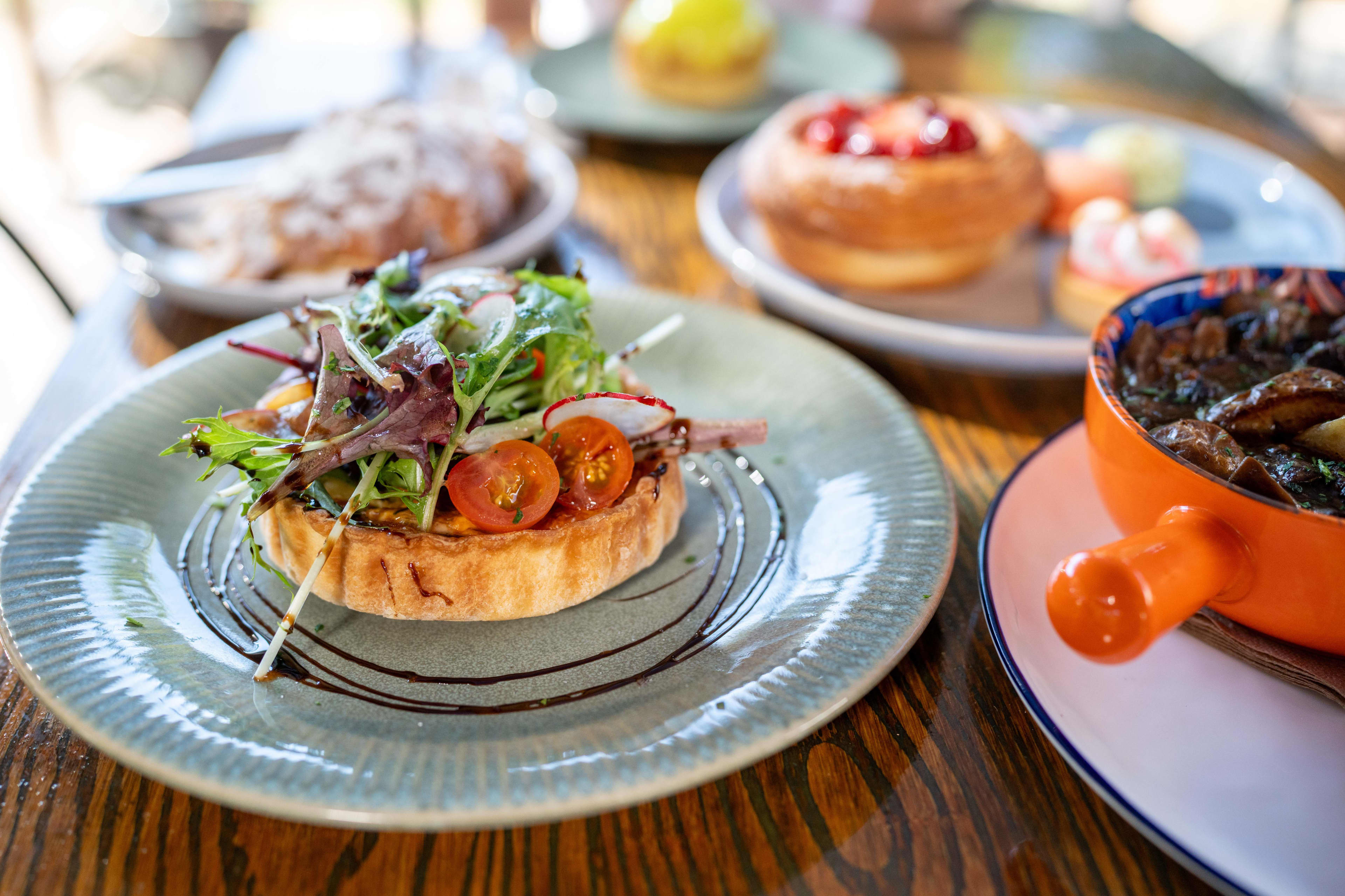 An array of food on a table, with a tart on a grey plate with tomatoes and greens on top in the foreground.