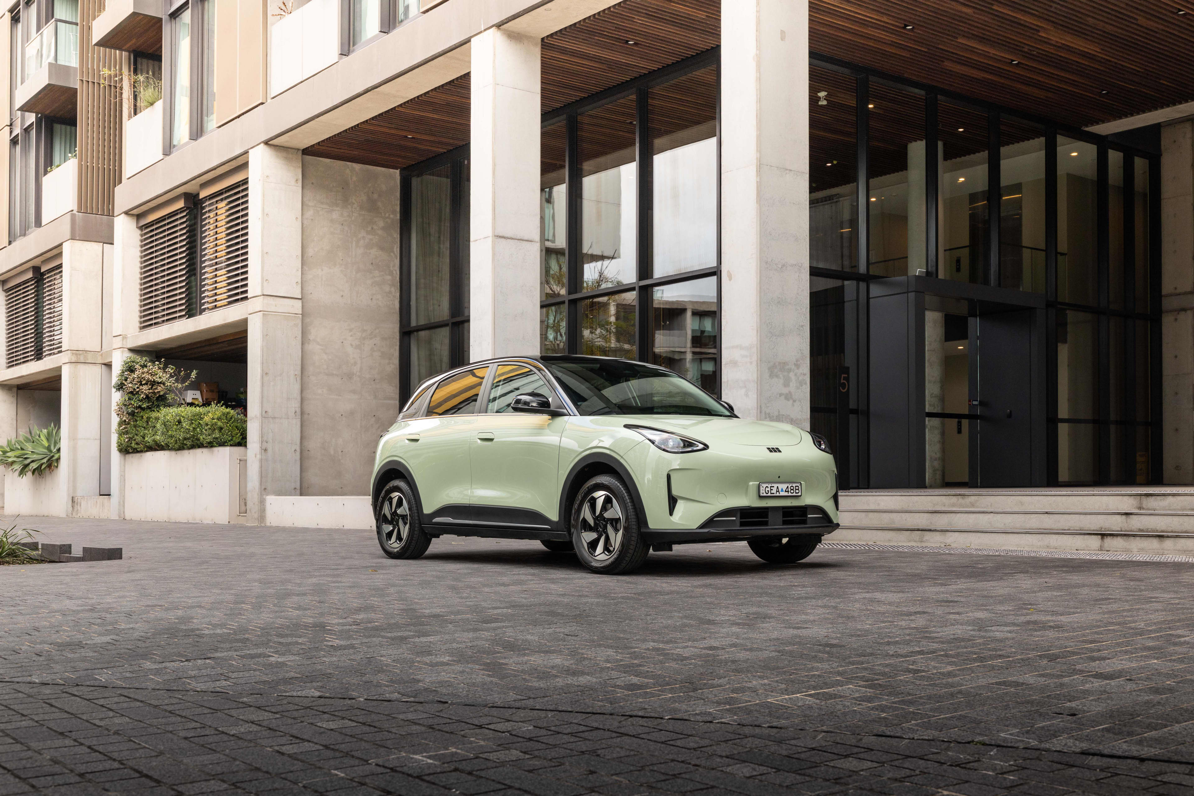 A mint green Geely EX2 Ev parked on dark coloured paving outside a city building with pale columns and dark glass.
