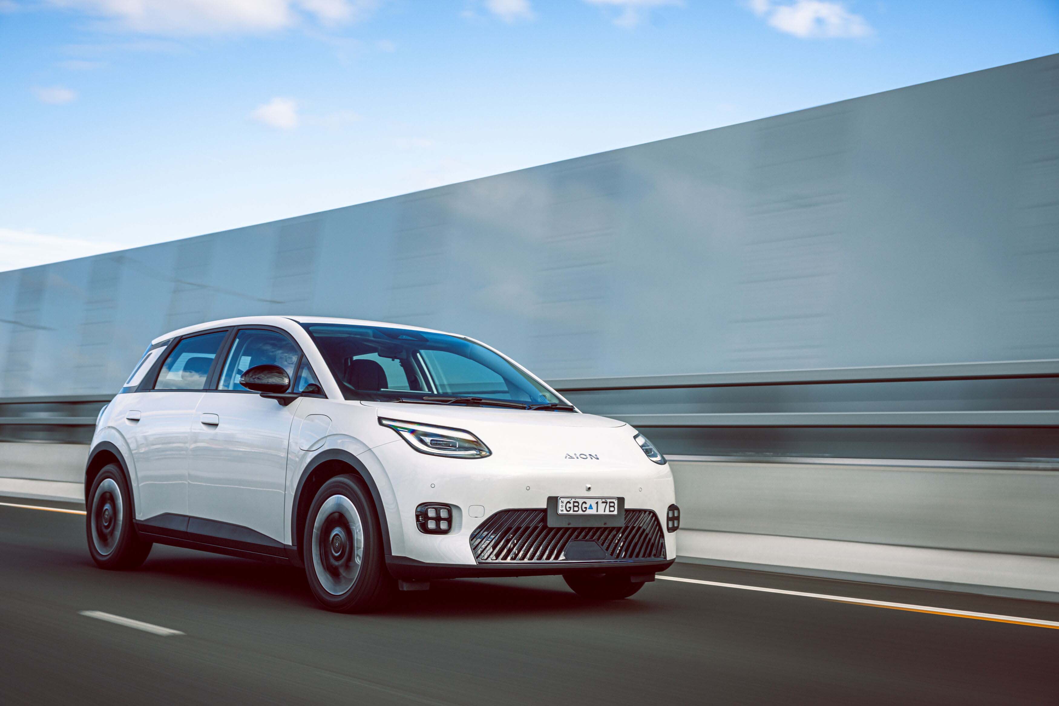 A white GAC Aion UT EV driving along a bitumen road, with a wall blurred in the background.