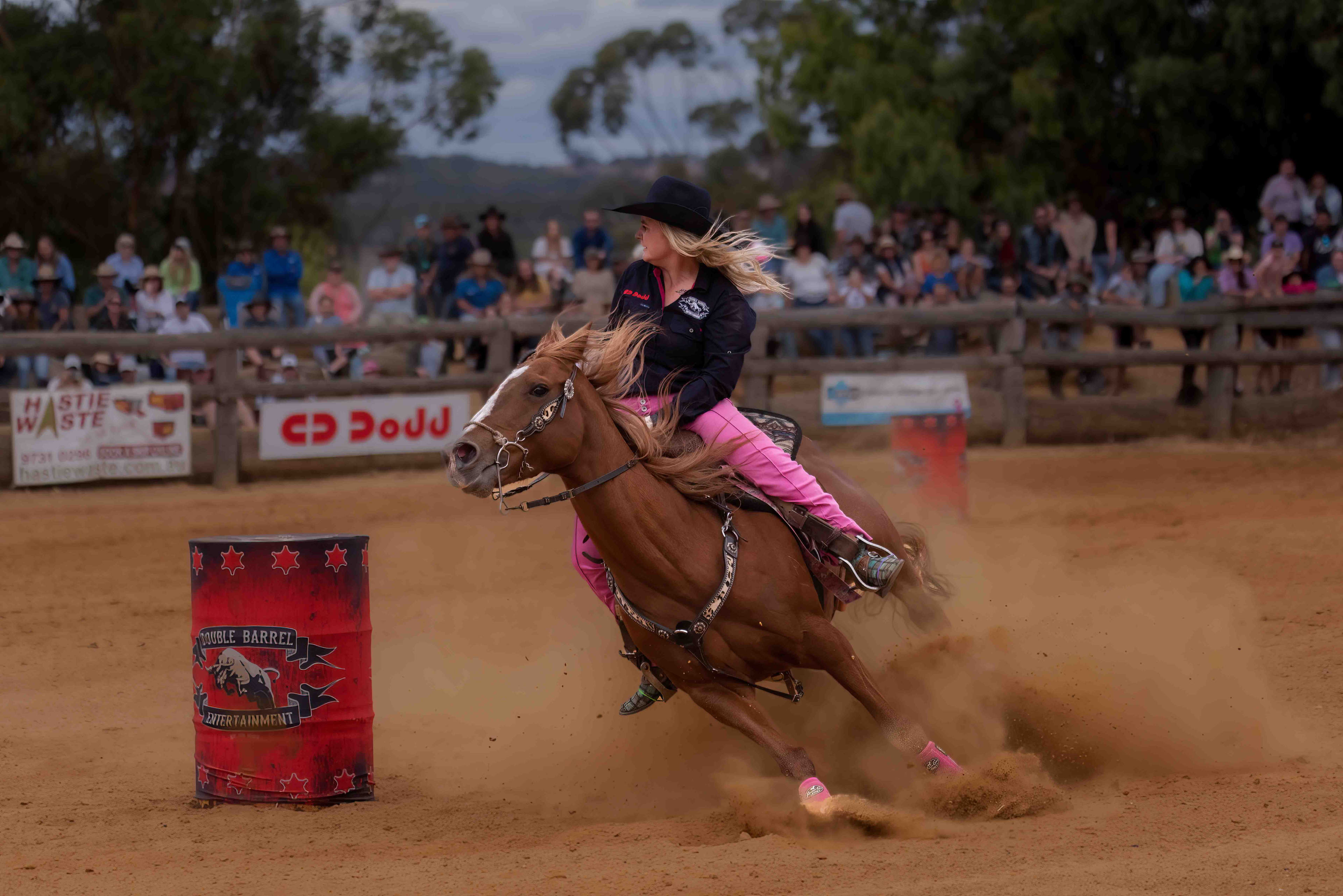 A lady with long blonde hair and a wide brimmed black hat riding a brown horse around a barrel that reads "Double Barrel Entertainment".