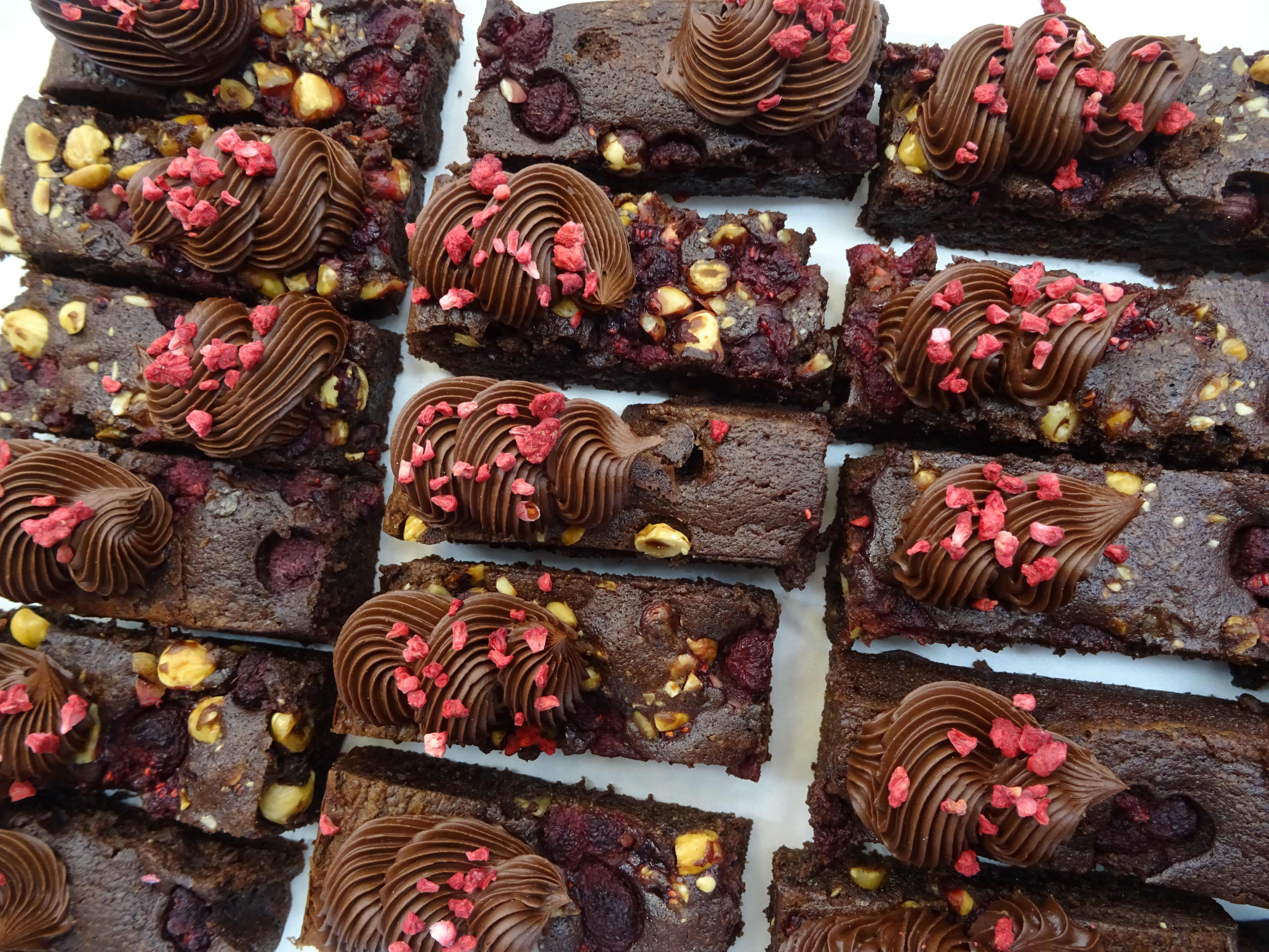 A top-down view of several rectangular chocolate brownies arranged in rows. Each brownie is topped with a piped swirl of chocolate frosting, chopped nuts, and sprinkles of freeze-dried raspberry pieces.