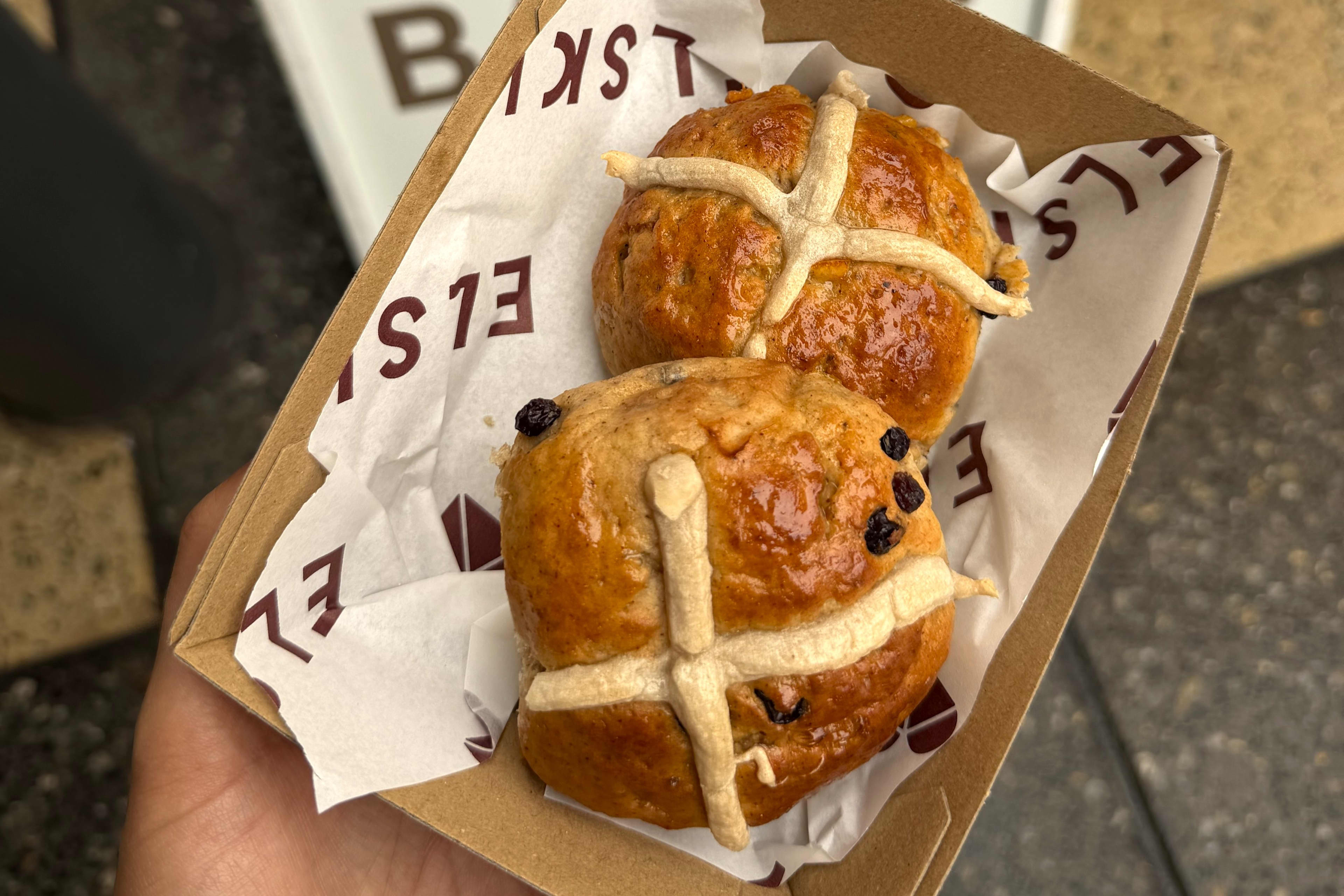 Two golden hot cross buns sitting in a cardboard tray with "Elski" written in brown writing.