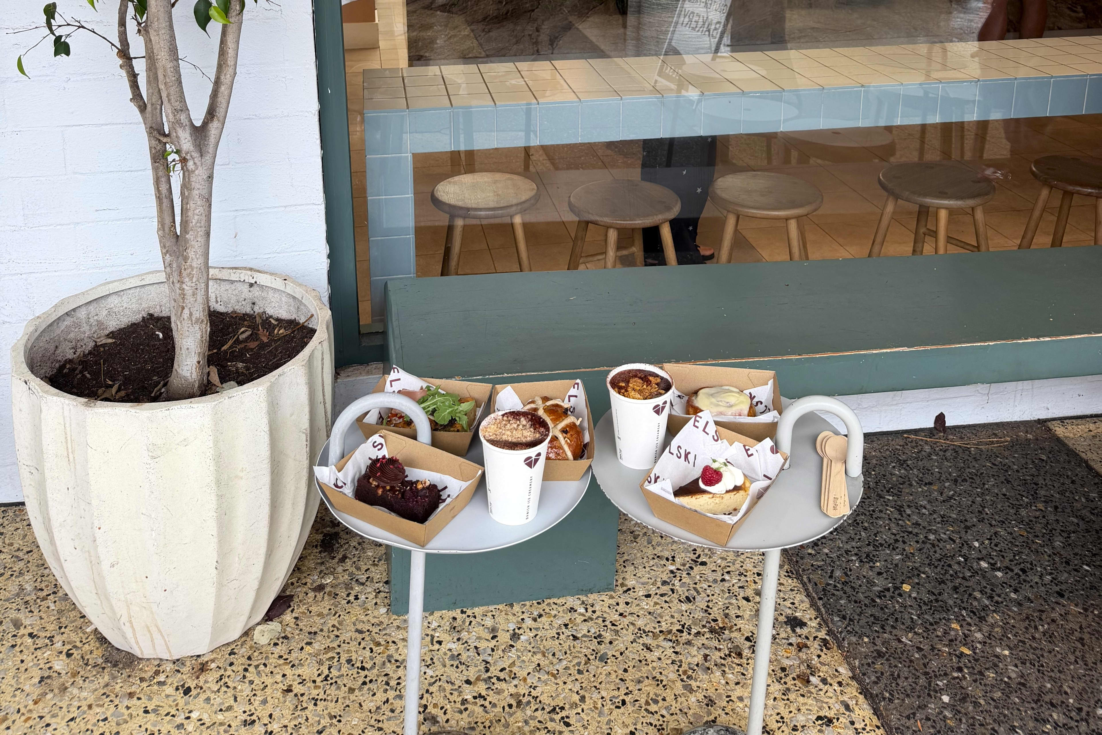 Two small, round white outdoor table stand side-by-side, holding an assortment of cafe food and drinks.