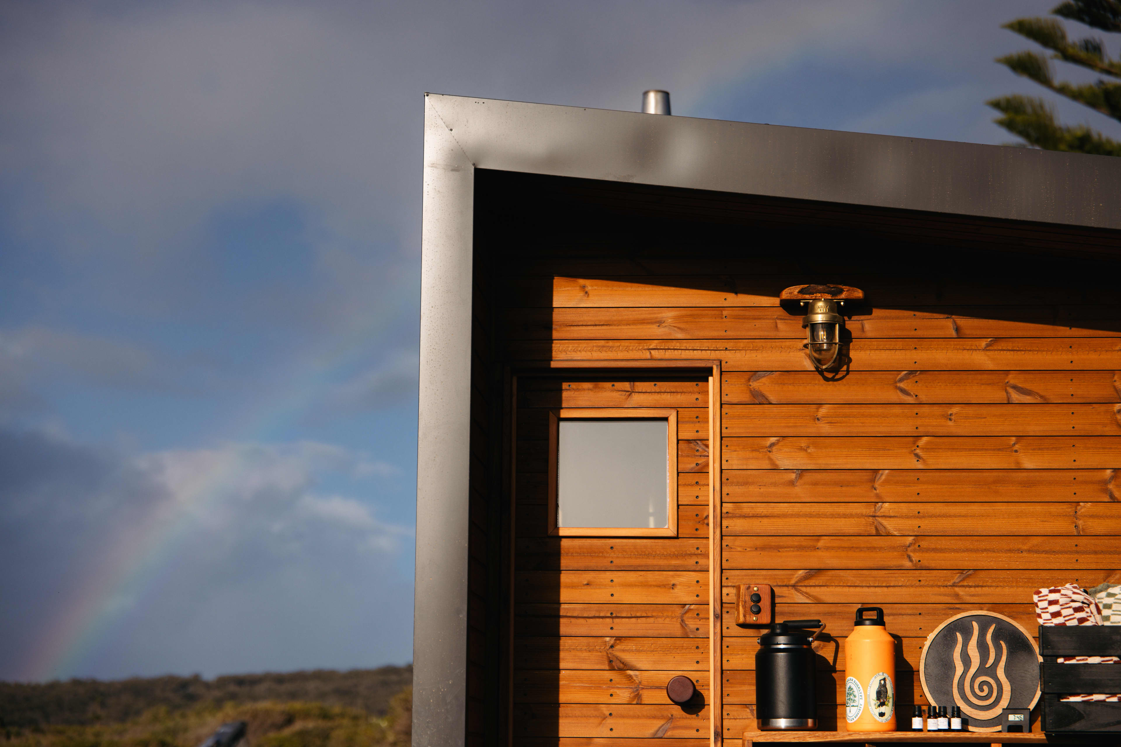 Rainbow in background of small timber sauna with water bottles outside.