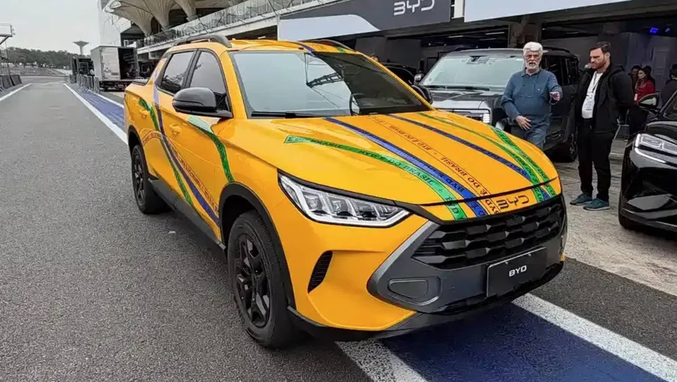 BYD's Mako ute in yellow, with green adn blue stripes, parked on the side of a track, with two men looking on.