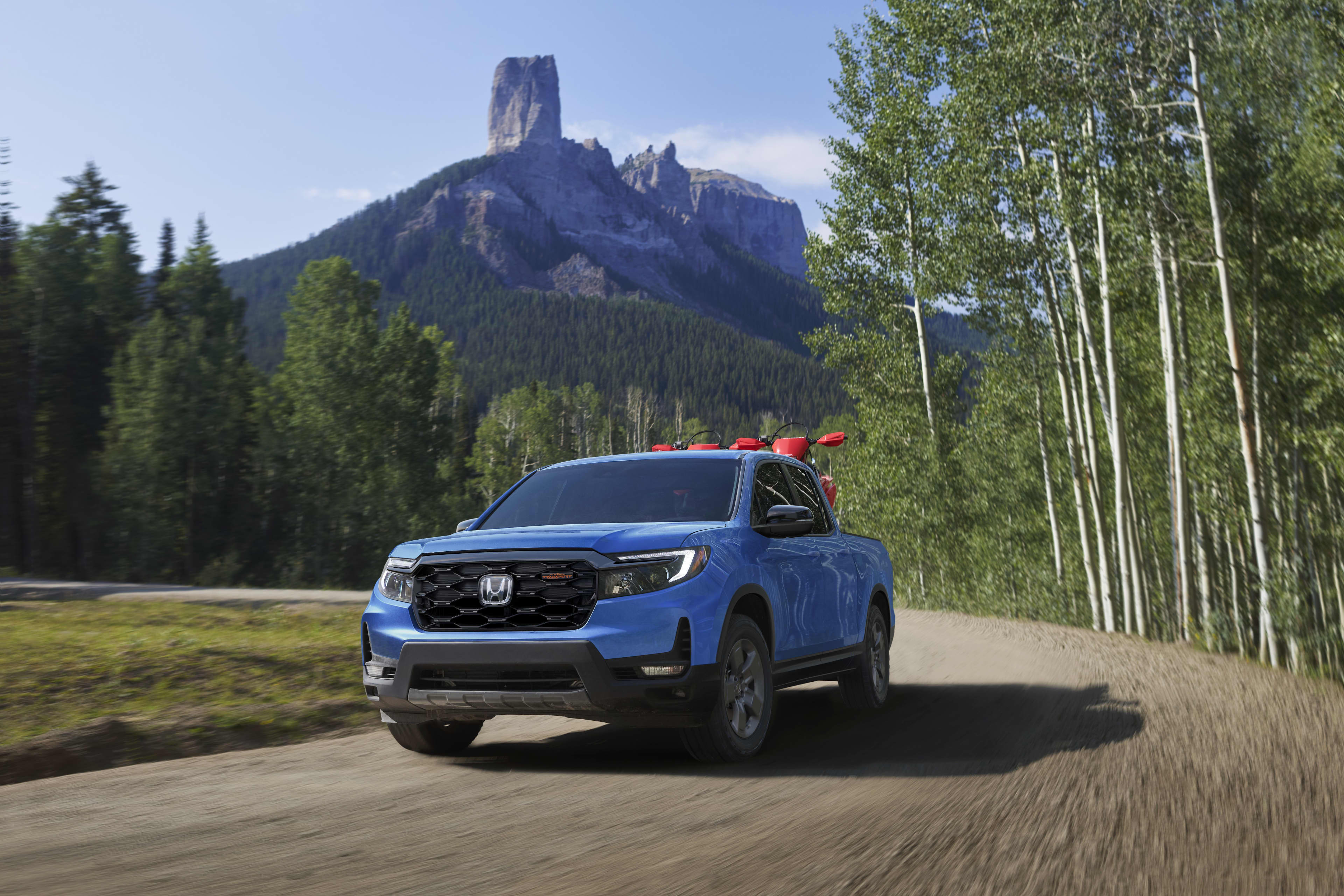 A blue Honda Ridgeline Trailsport ute driving on a dirt road, with trees and a mountain in the background.