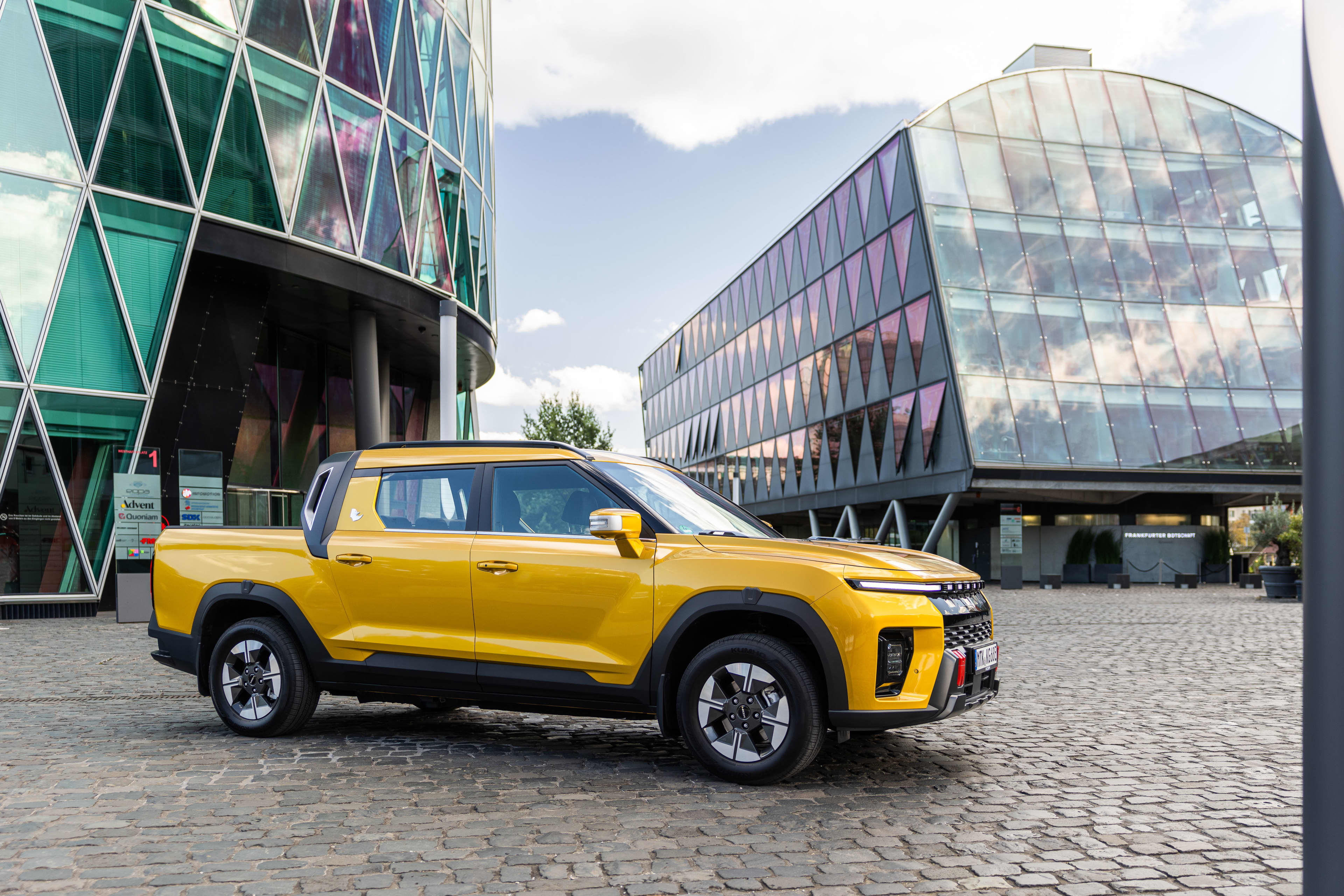 A yellow KGM Musso EV ute with glass city buildings in the background.