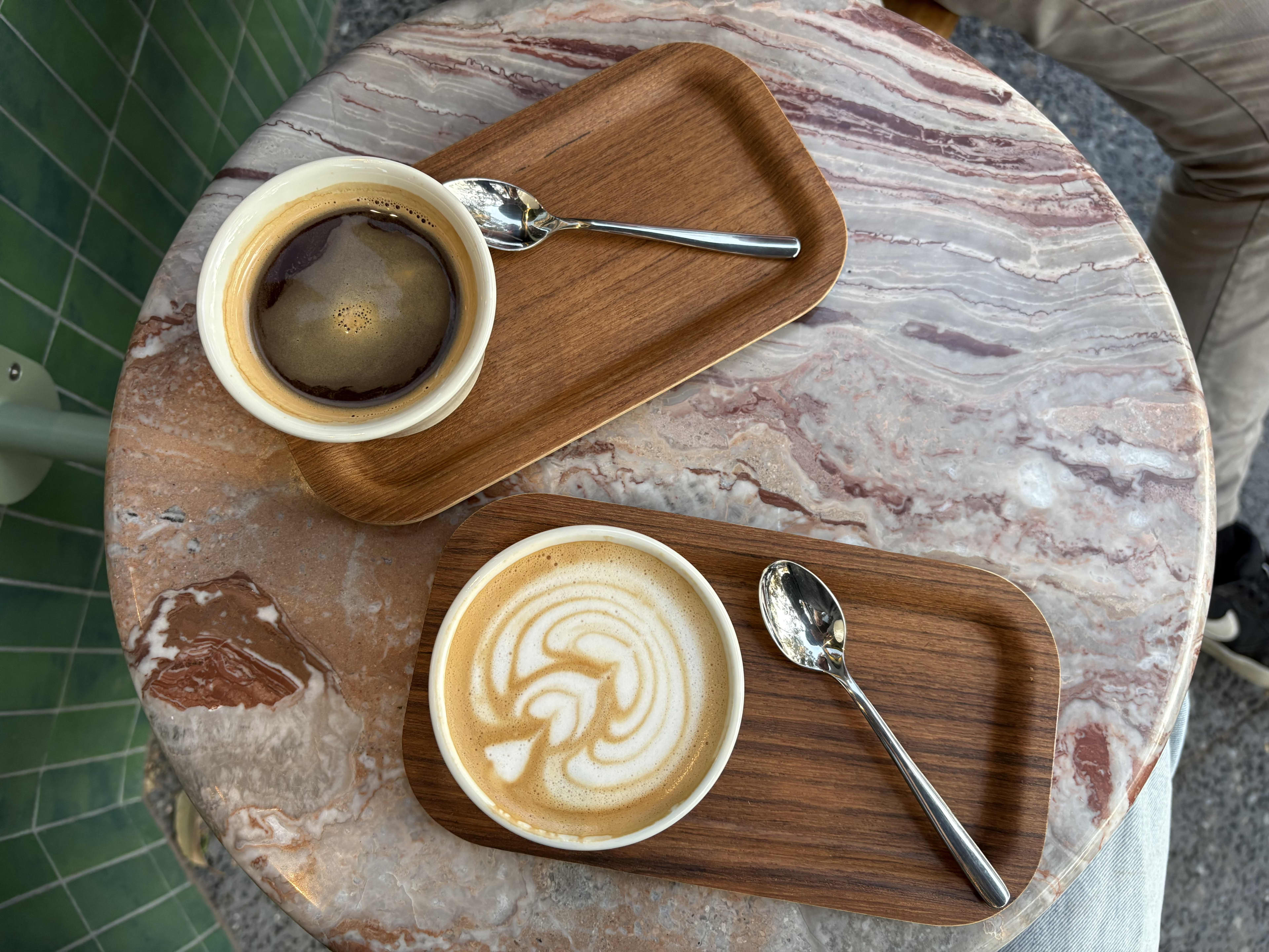 An overhead shot shows two cups of coffee served on individual small wooden trays atop a pink and white marbled table.