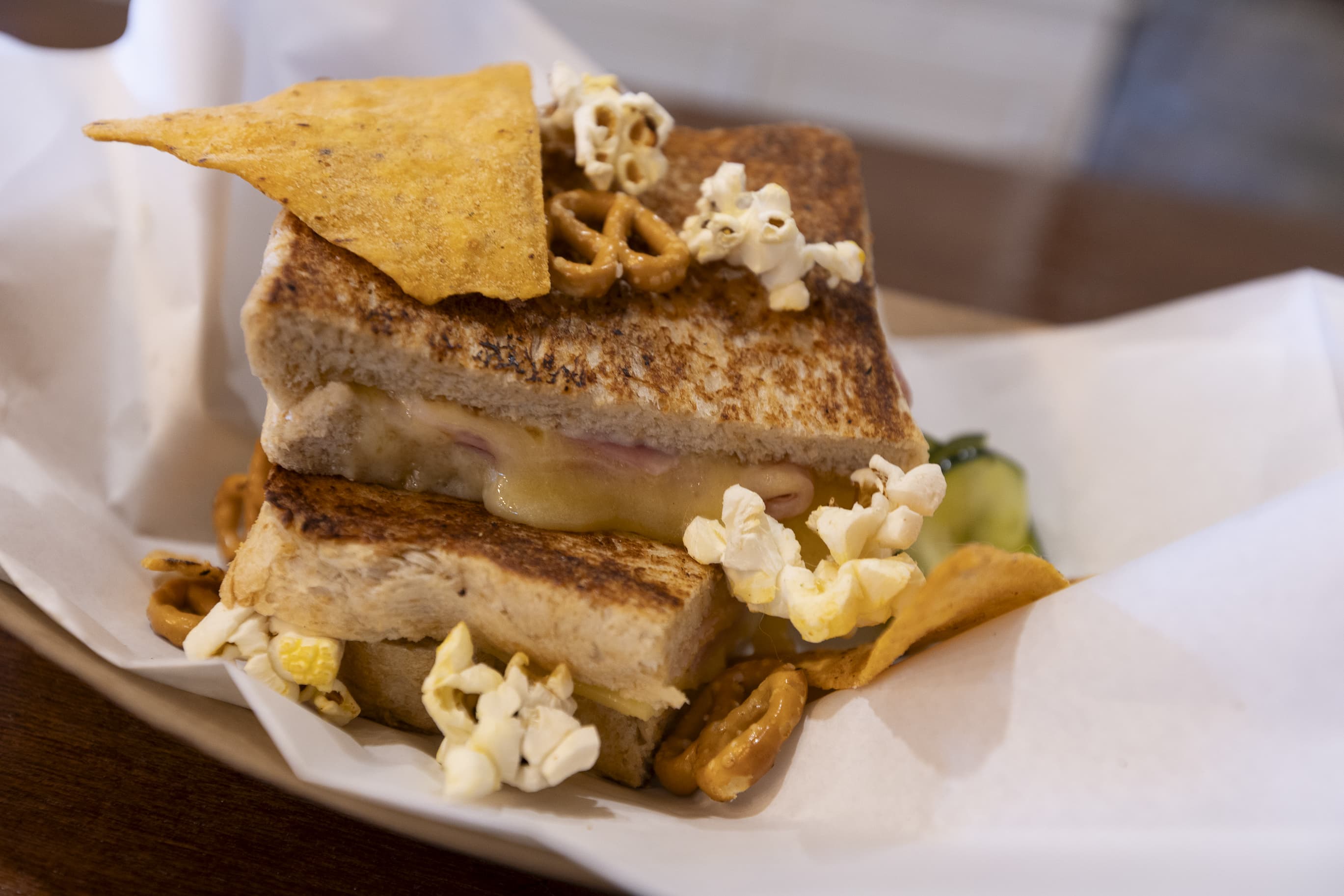 A toasted sandwich sitting in white paper filled with ham and cheese and topped with popcorn, pretzels and a corn chip.