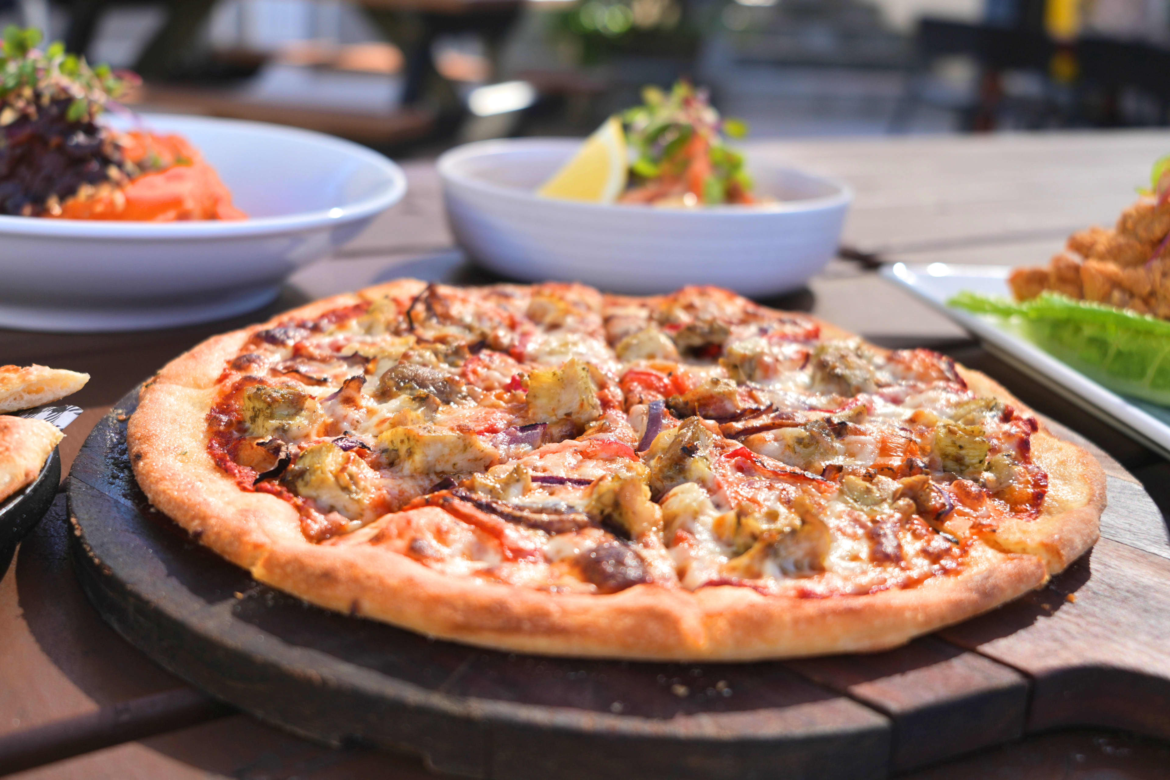 A large tomato based pizza topped with chicken and onion, with meals placed in the background.
