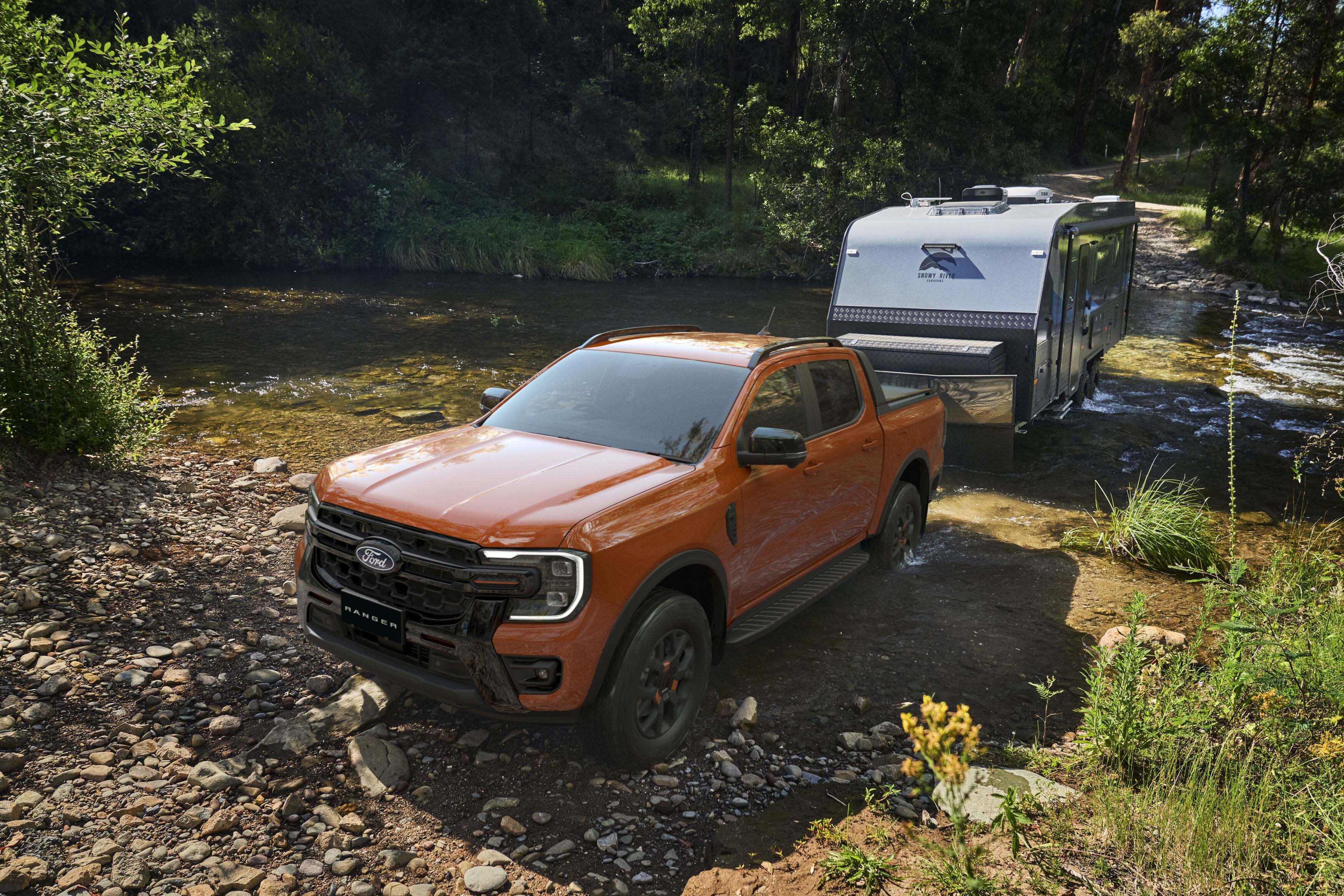 An orange Ford Ranger ute towing a caravan through a creek in the forest.