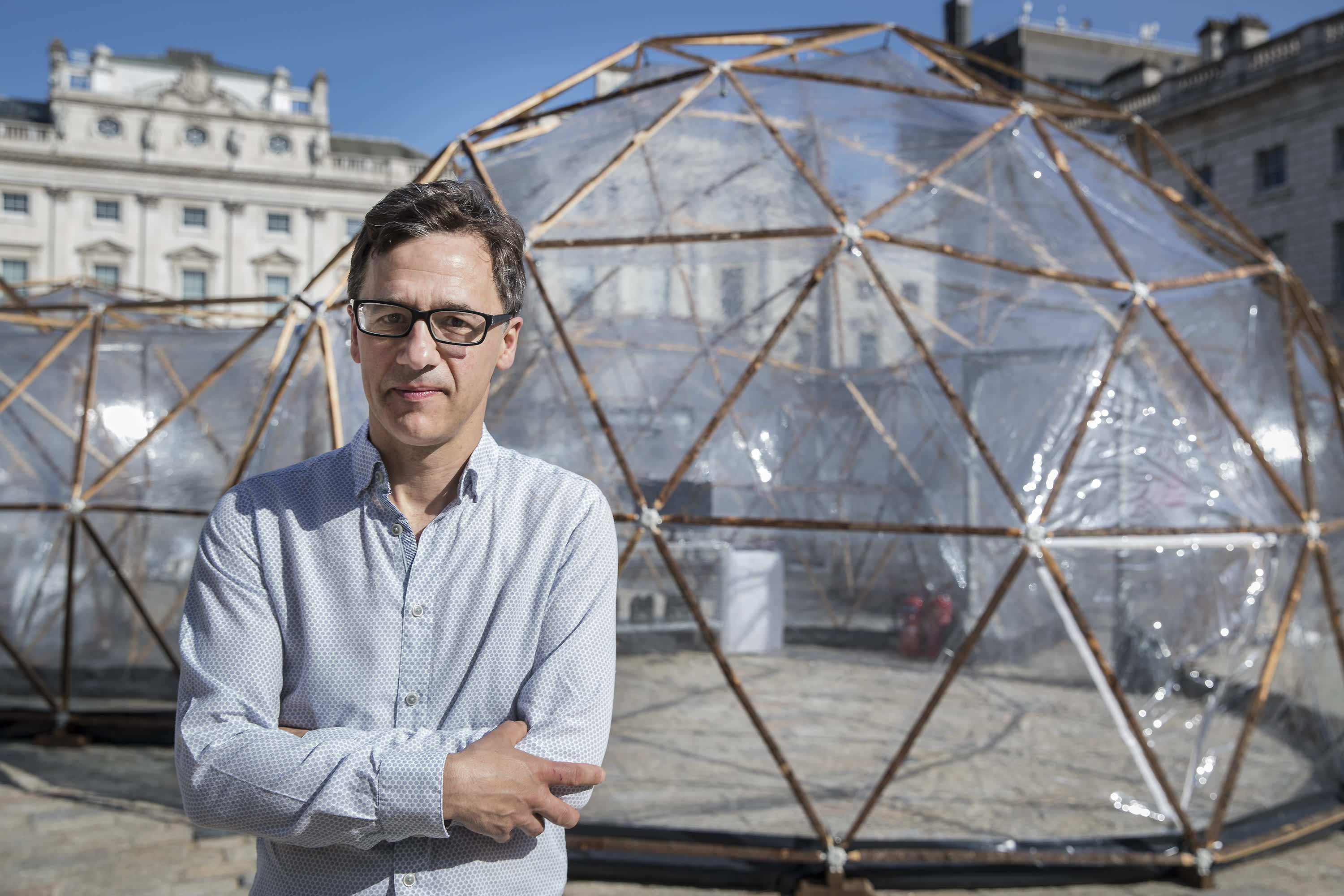 Artist Michael Pinksy stands with his arms crossed in front of a framed transparent dome, as part of a previous installation of his Pollution Pods exhibition.