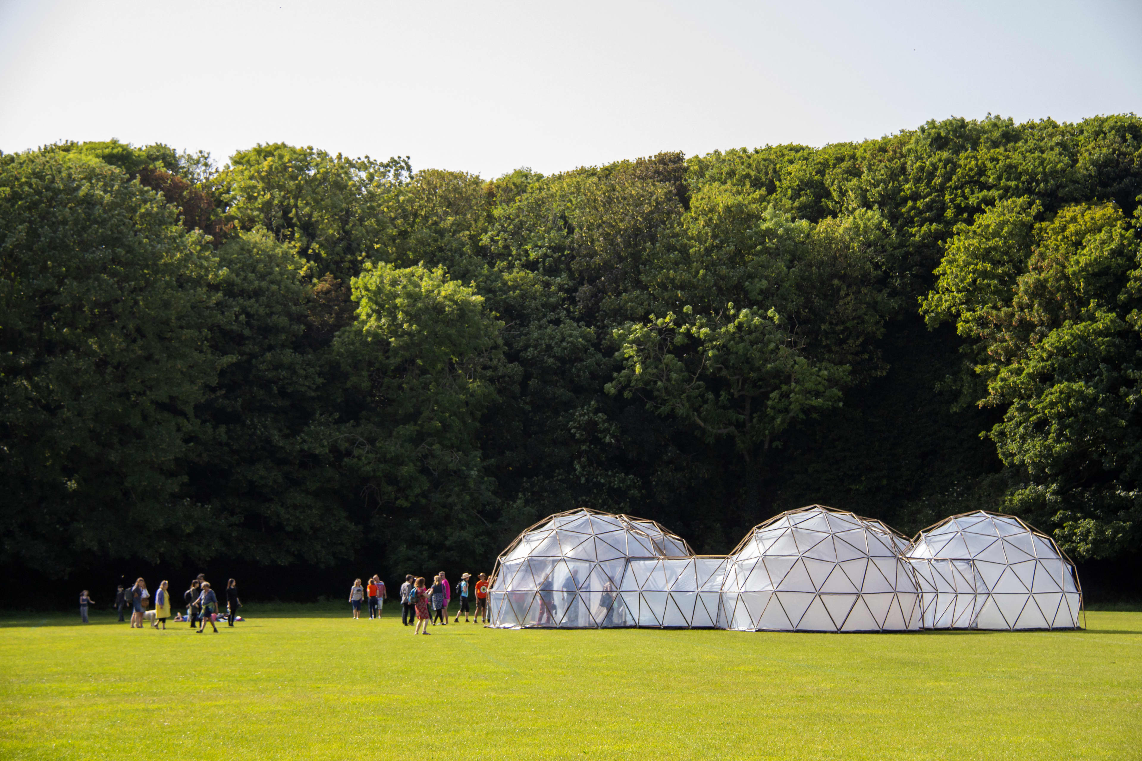 A collection of framed partially transparent domes on grass in front of a tall stand of trees, with people standing nearby, as part of a previous installation of artist Michael Pinsky's Pollution Pods. Photo credit: Rachel Dunford.