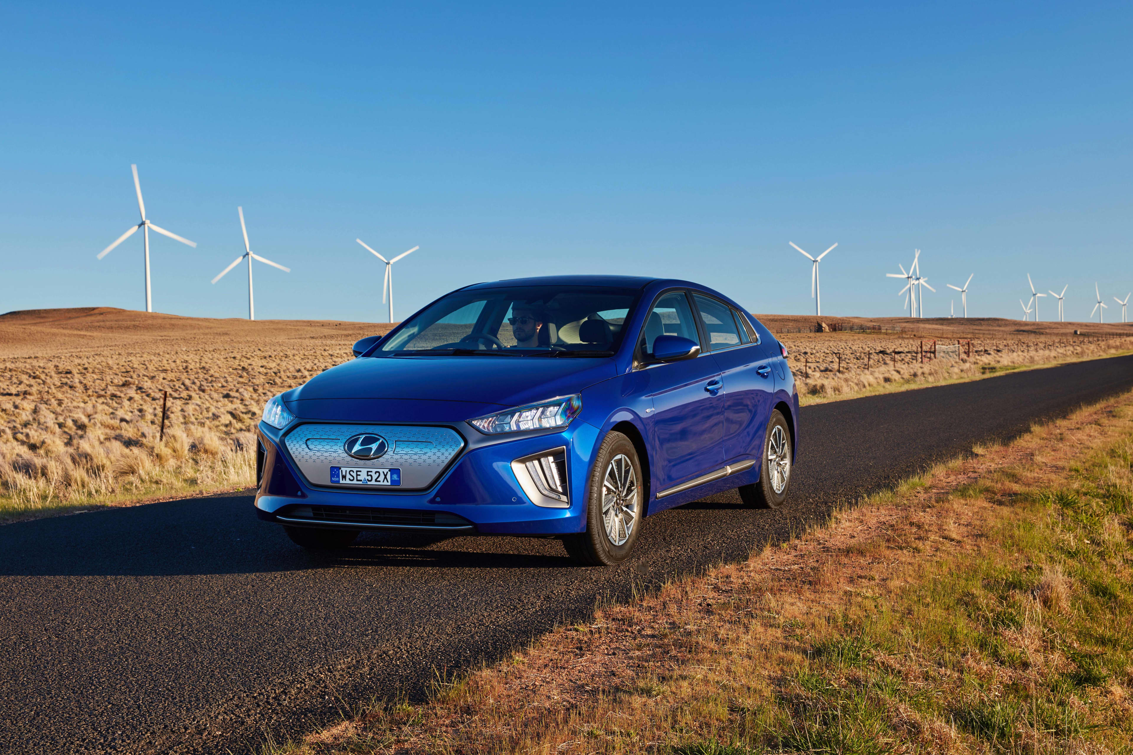 A blue 2019-2022 Hyundai Ionic EV drives on a bitumen road through a landscape of dry grass, with wind power turbines in the background against a blue sky.